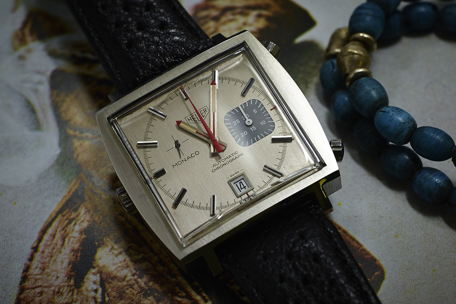 A close-up of the Heuer Monaco 1533G chronograph with a square silver case, cream dial, black sub-dial, and date window on a black leather strap. Blue beaded bracelets are beside this vintage Heuer wristwatch.