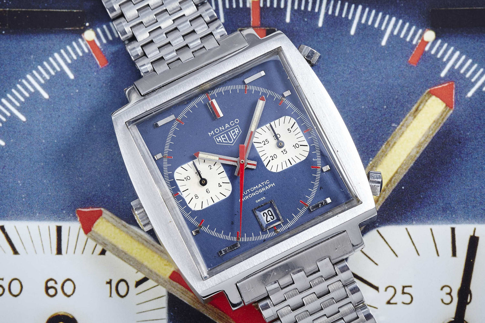 A square-faced Heuer Monaco watch with a blue dial and stainless steel bracelet is set against bold, colorful graphics—a true tribute to Steve McQueen’s iconic style.