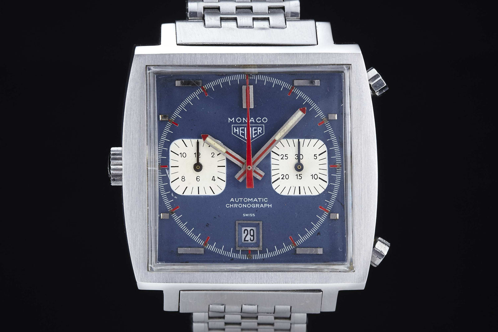 A close-up of the Heuer Monaco by Heuer, featuring a square stainless steel case, blue dial with two white subdials, date at 6 o’clock, and metal bracelet—set against a black background.