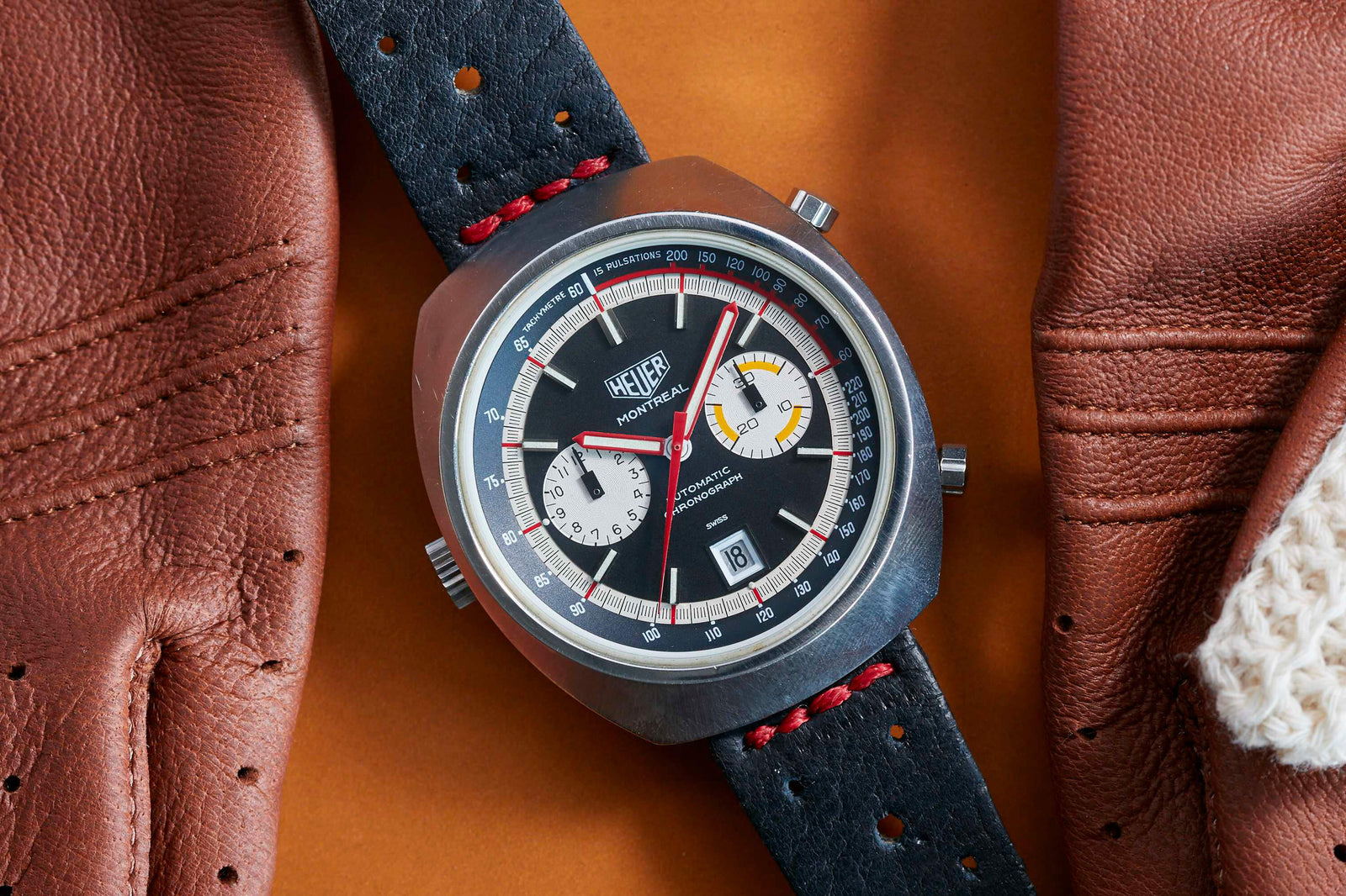A vintage Heuer Montreal automatic chronograph with a black dial, two subdials, and date window is shown on a black perforated leather strap with red stitching, nestled between brown leather driving gloves.
