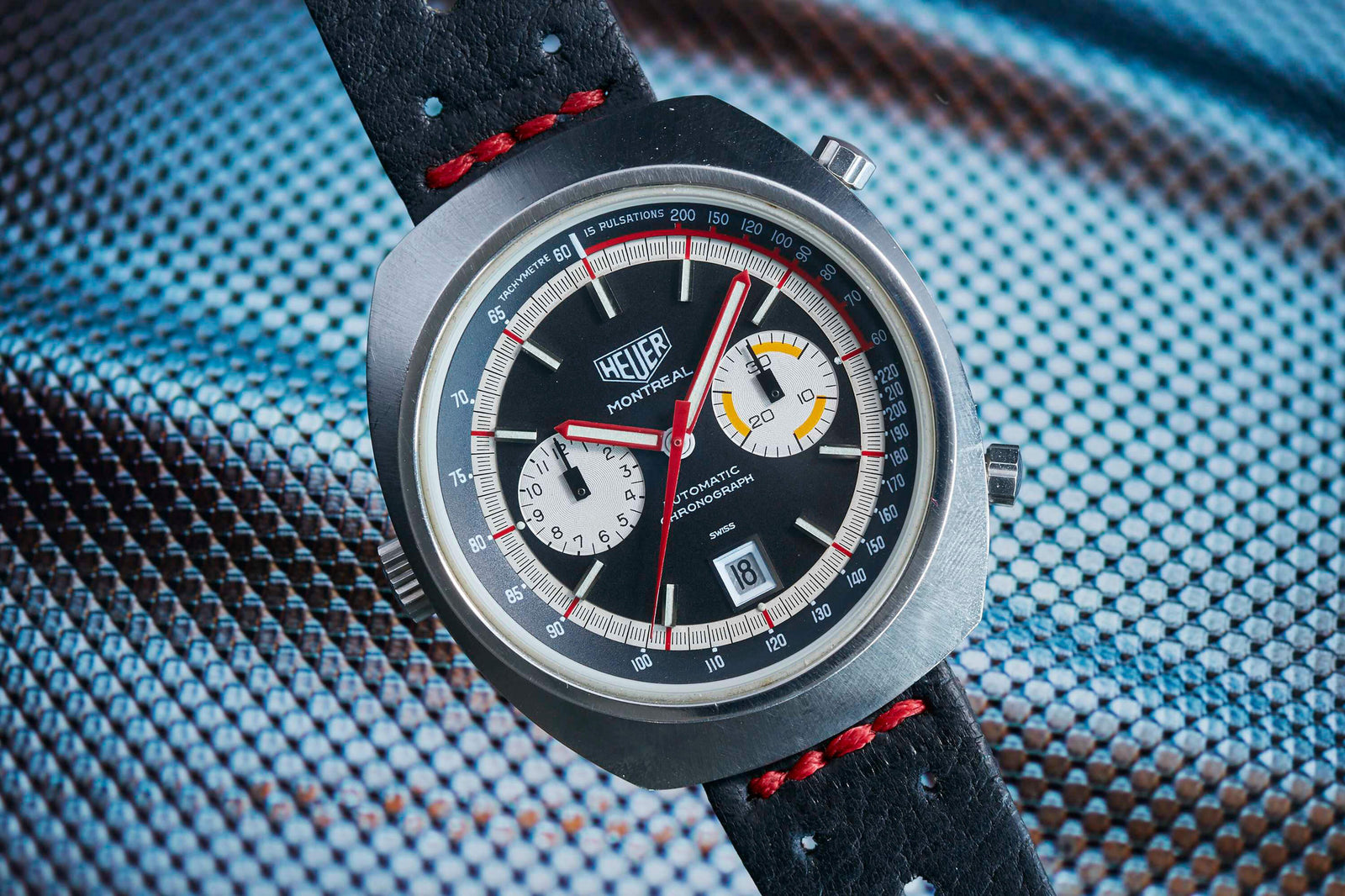 A close-up of the Heuer Montreal by Heuer, featuring a black dial with red and yellow accents, three subdials, a date window, and a black leather strap with red stitching on a textured metallic surface.