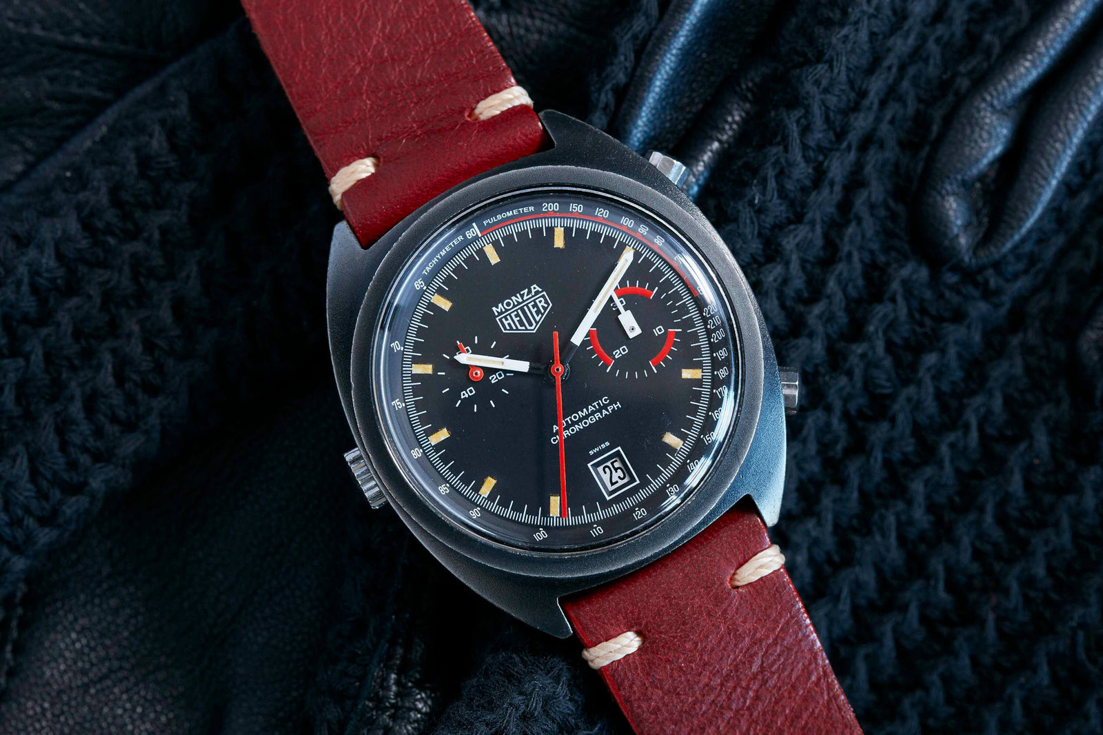 A vintage Heuer Monza wristwatch with a black dial, red and white hands, chronograph subdials, and a date display on a red leather strap—highlighting Heuer’s Formula 1 racing heritage—rests on a textured black surface.