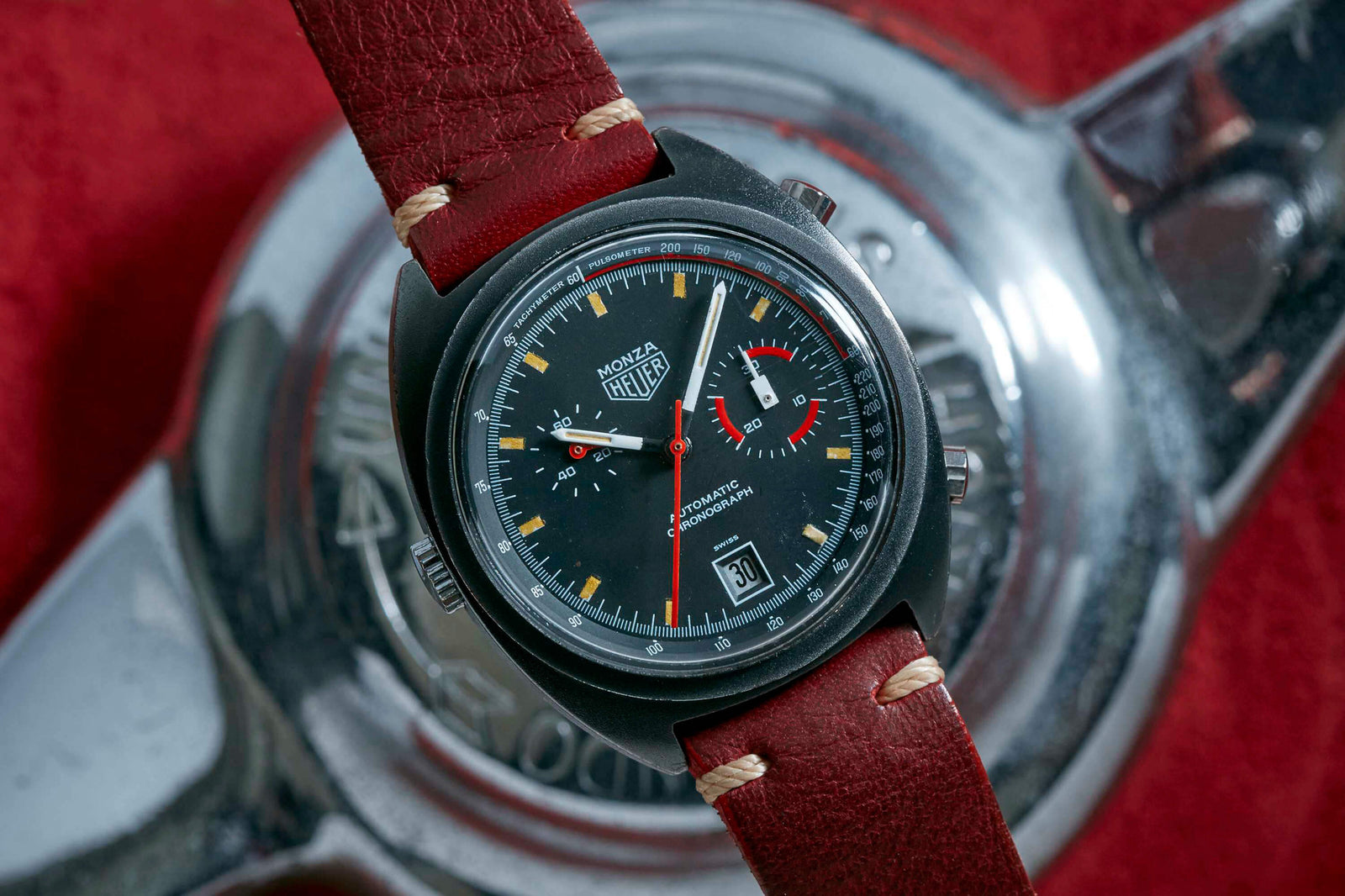 A vintage Heuer Monza wristwatch with a black dial, red accents, and date display sits on a shiny metallic car wheel part, reflecting Heuer’s Formula 1 heritage. The brown leather strap with beige stitching pops against the red background.