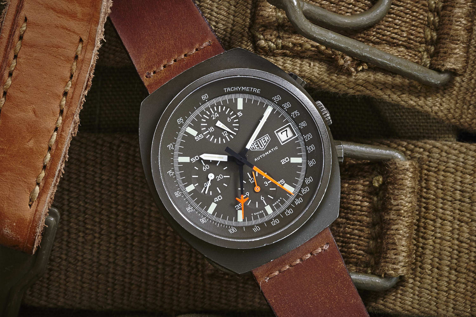 A Heuer Lemania Chronograph by Heuer with a brown leather strap rests on olive green fabric with metal buckles. The black dial showcases multiple subdials, white markings, and vibrant orange hands.
