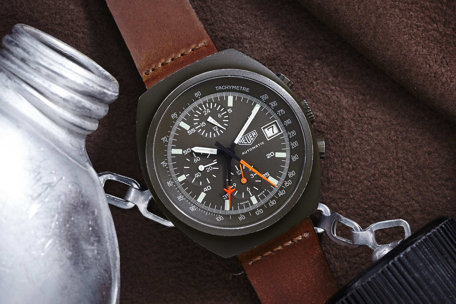 A Heuer Lemania Chronograph with a brown leather strap rests on a brown fabric surface beside a metal canteen and silver chain. This vintage-inspired piece features a black face, white markers, subdials, and orange accents.
