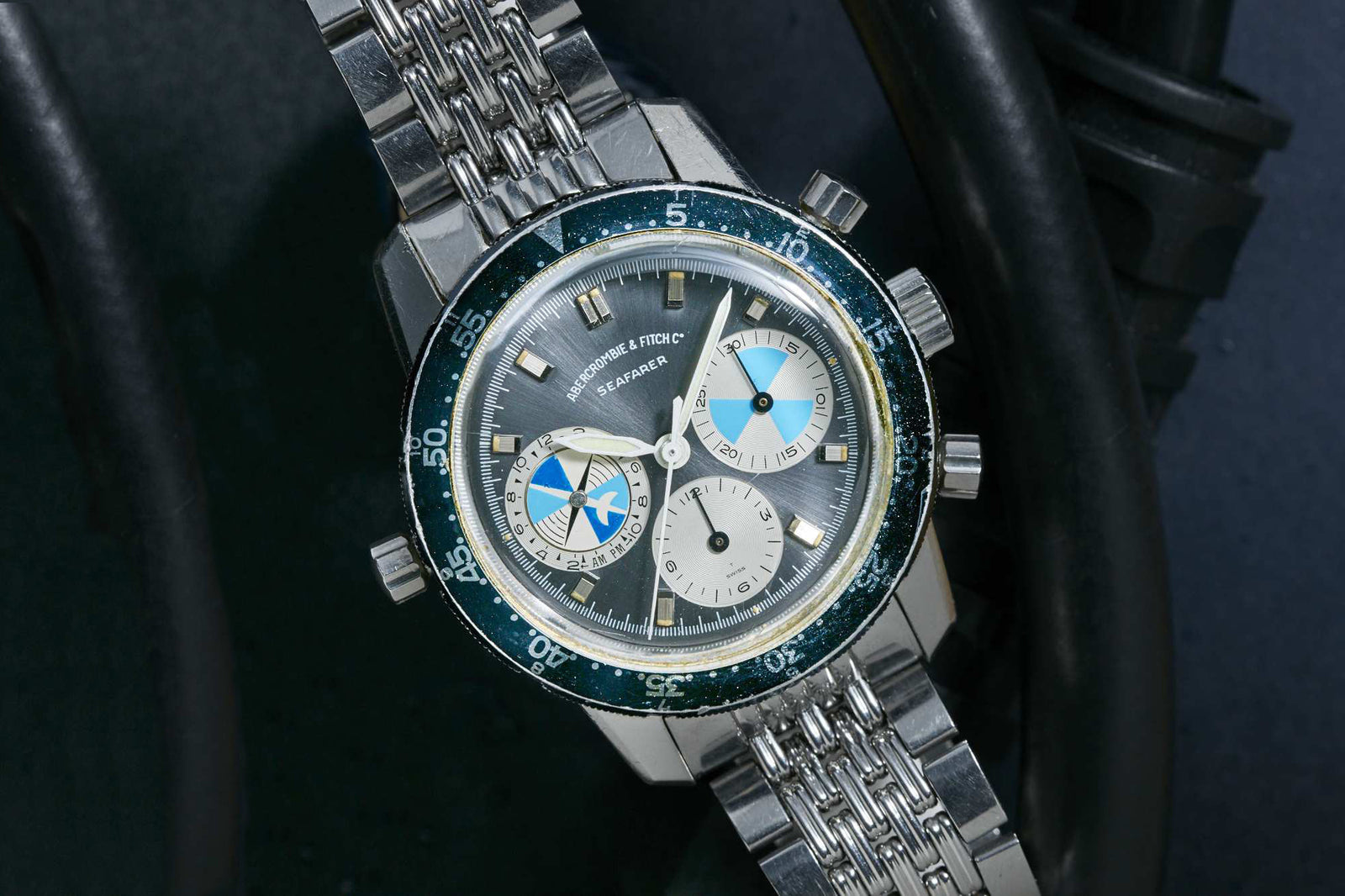 The Heuer Seafarer for Abercrombie & Fitch features a silver bracelet, blue and silver bezel, and two subdials—one with a blue and white design—resting on a dark surface among black cables. Detailed description coming soon.