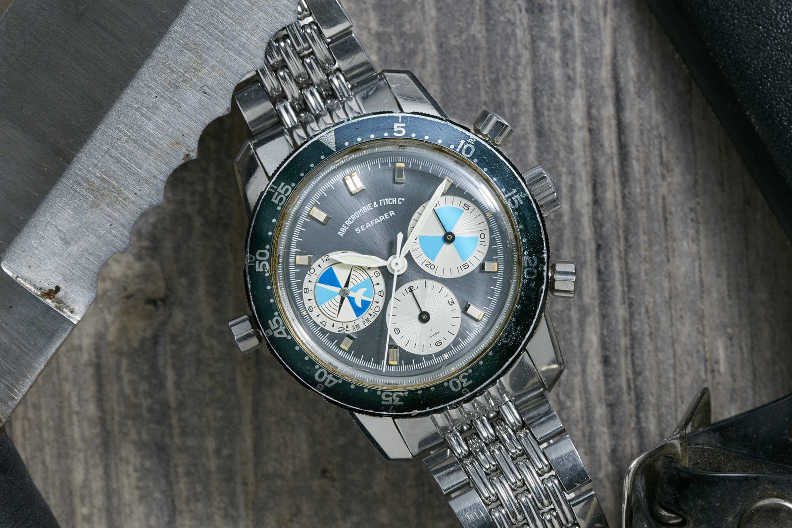 A Heuer Seafarer for Abercrombie & Fitch vintage silver wristwatch with a blue and white dial, three subdials, and a metal link band rests on a wooden surface beside a serrated metal object. Detailed description coming soon.