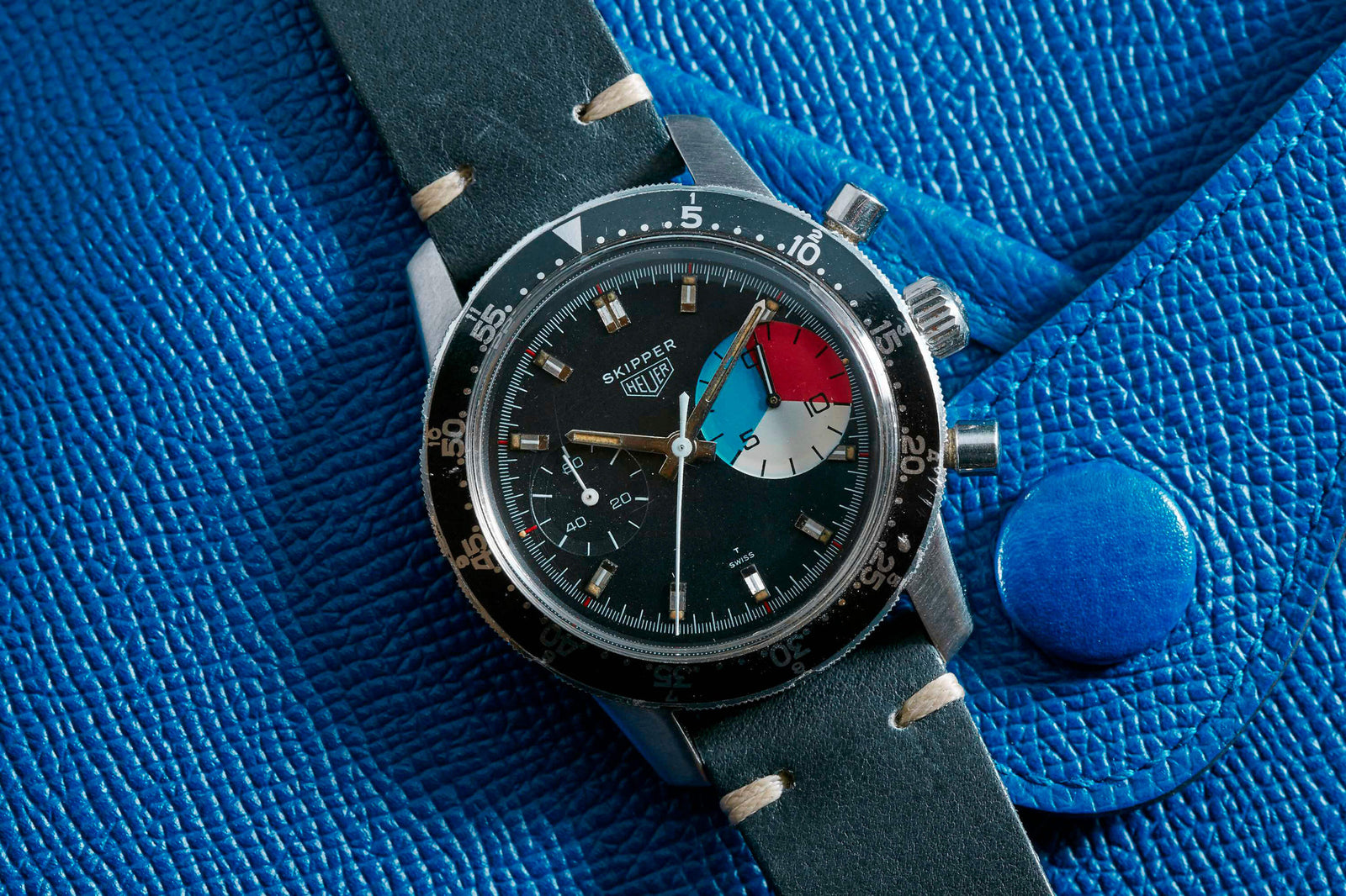 A Heuer Skipper regatta timer by Heuer, featuring a black dial, colorful sub-dial, and black leather strap, rests on a textured blue leather surface with a large button.