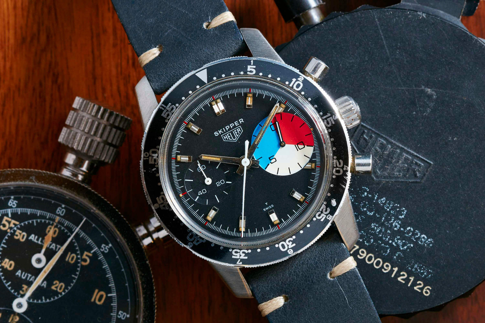 A close-up of the Heuer Skipper by Heuer, featuring a black dial with colorful red, blue, and white subdial, silver markers, and a black leather strap. The watch is placed next to another stopwatch on a wooden surface.