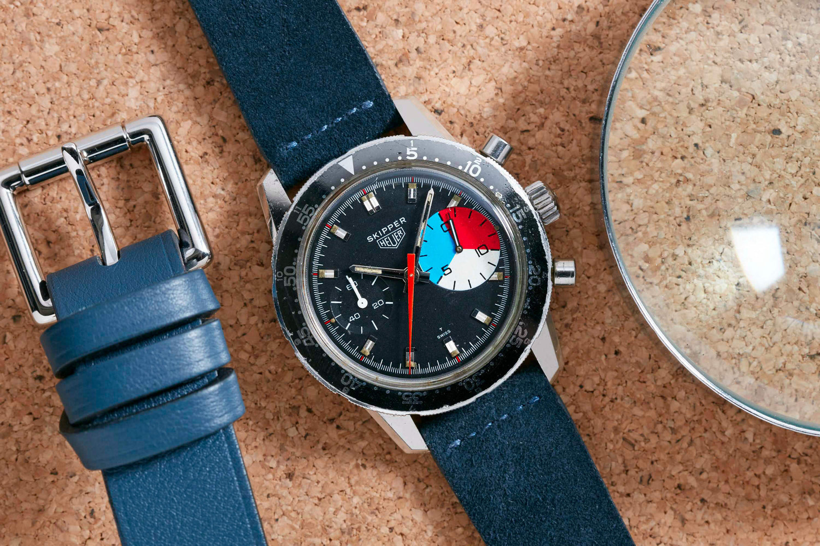 A vintage Heuer Skipper Reference 7764 chronograph watch with a blue strap, black dial, and colorful subdial rests on a cork surface beside a matching belt and magnifying glass. Detailed Description Coming Soon!.