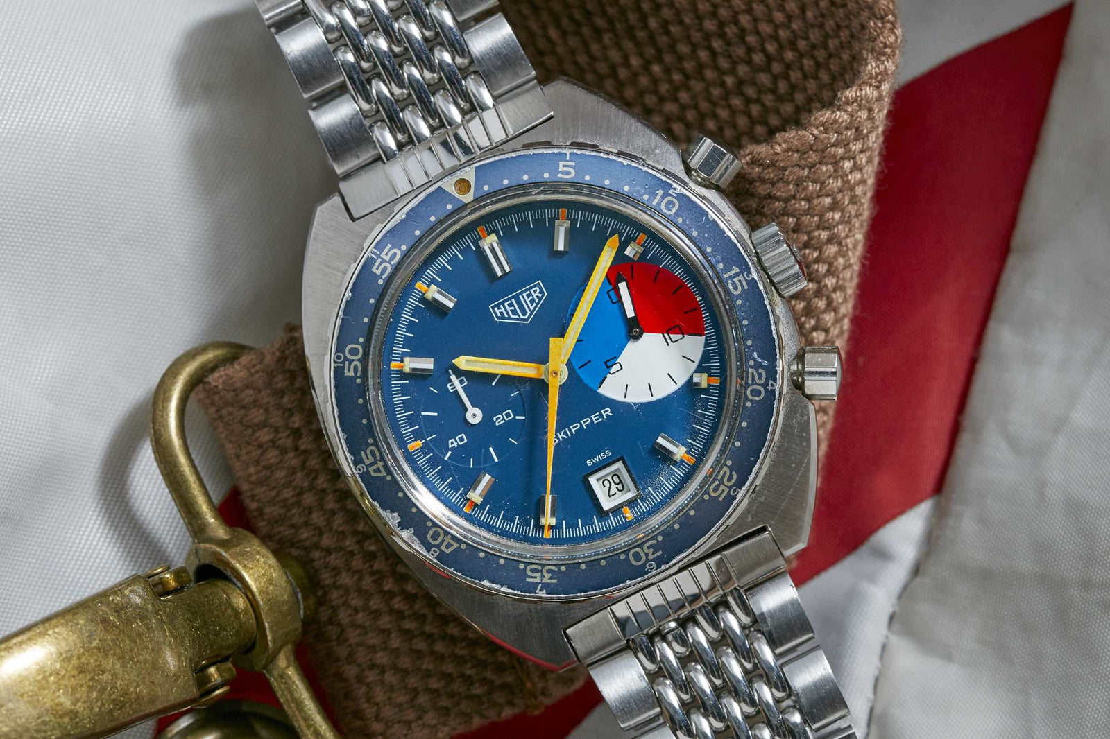 A Heuer Skipper Manual Wind watch with a blue dial, multicolor subdial, and silver case is presented on a brown strap against red and white fabric—a classic sports timepiece by Heuer.