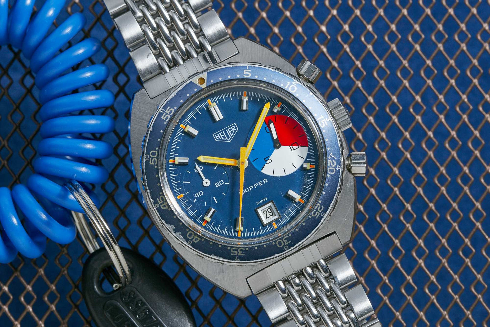A close-up of the Heuer Skipper Manual Wind by Heuer, featuring a blue dial, colorful subdial, and metal bracelet on a blue mesh background beside a coiled blue keychain and black key.