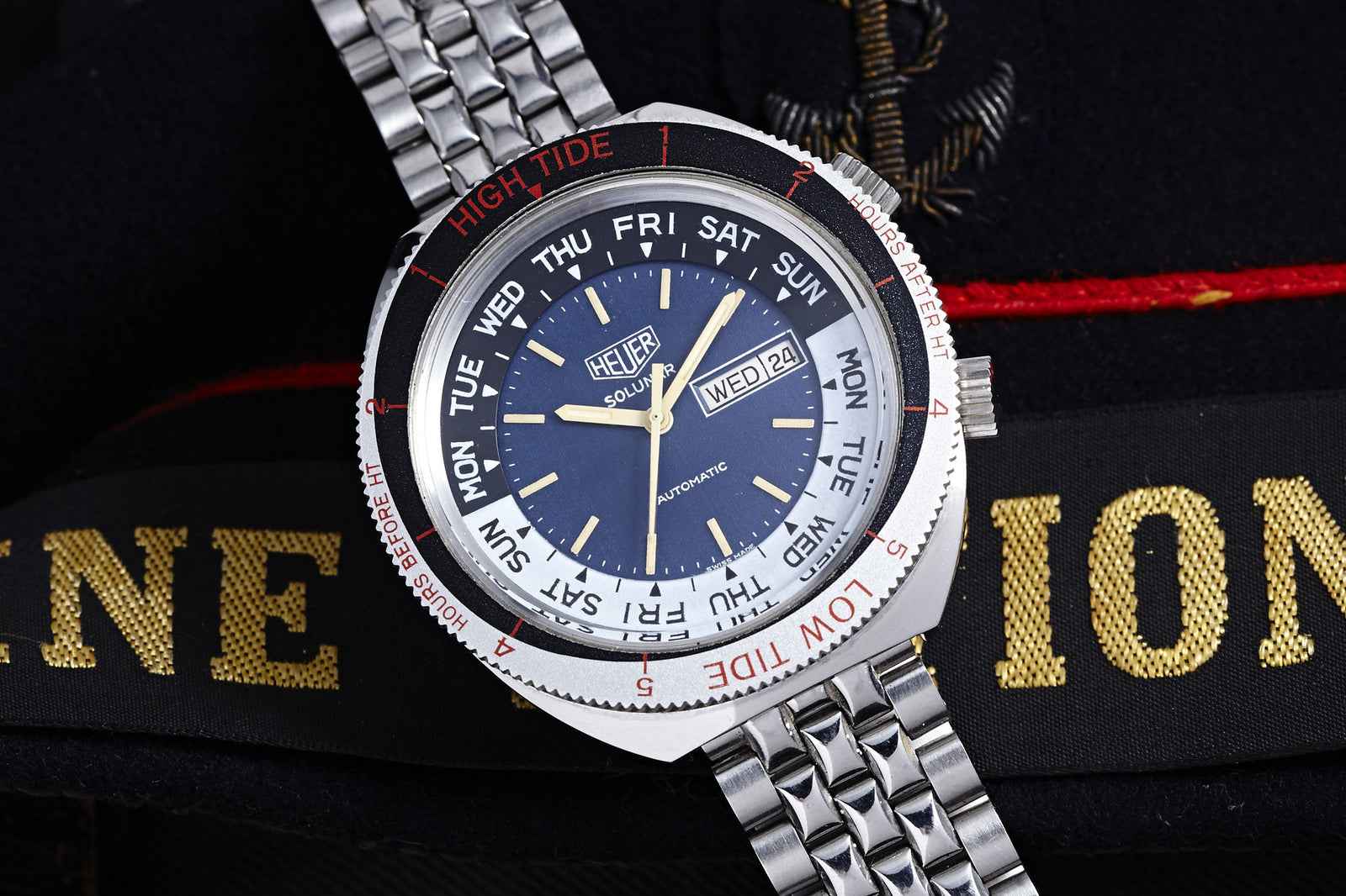 A silver Heuer Solunar Automatic with a blue dial showing day and tide levels rests on dark, gold-embroidered fabric with a red stripe—a modern interpretation of vintage Heuer watches.