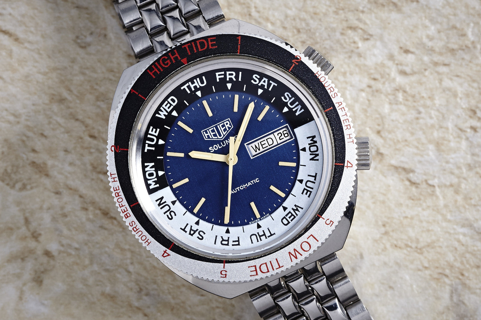 The Heuer Solunar Automatic by Heuer features a stainless steel band, blue and white dial, black day/date display, and a rotating bezel with HIGH TIDE and LOW TIDE in red on a textured beige surface.
