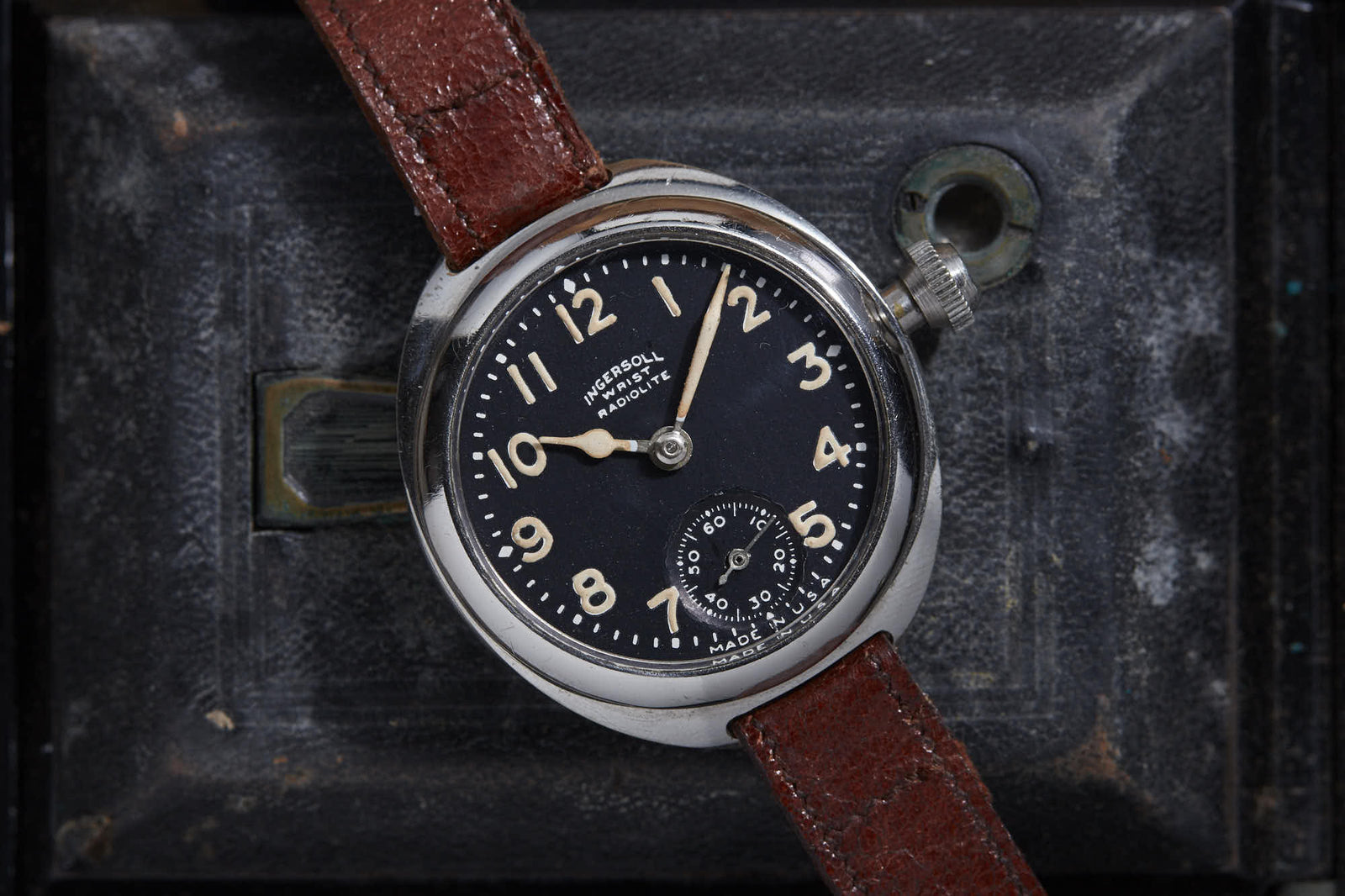 The Ingersoll Radiolite by Ingersoll, a vintage wristwatch with a brown leather strap and black dial with white numerals and small seconds subdial, lies on a worn black surface featuring a keyhole and metal latch.