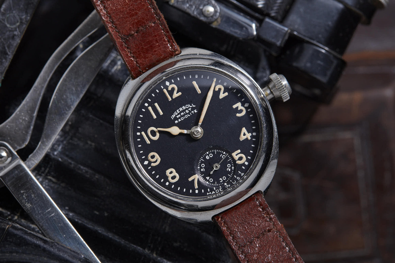 A close-up of the Ingersoll Radiolite wristwatch by Ingersoll shows its black dial, white Arabic numerals, and brown leather strap resting on a dark metal surface, reflecting vintage design inspired by World War I-era timepieces.