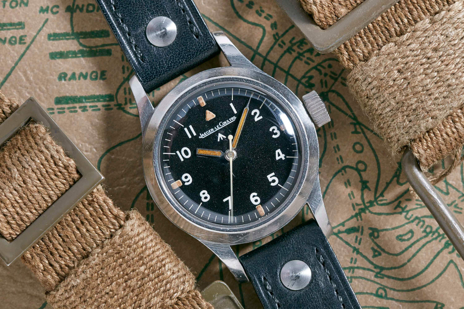 The Jaeger-LeCoultre JLC Mark XI Pilot’s Watch with a black dial, white numerals, and a black leather strap is shown on a brown map beside canvas watch straps.