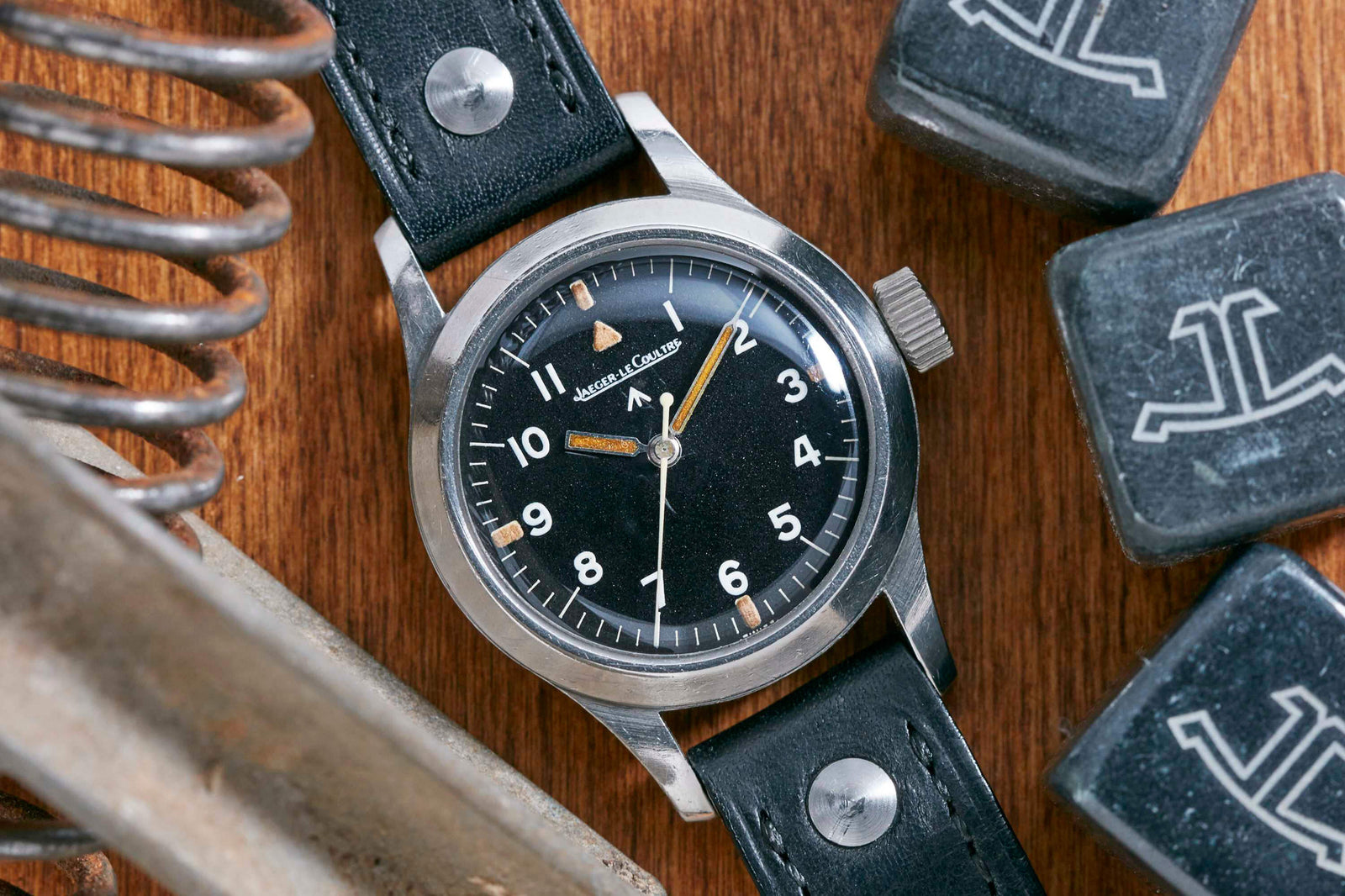 A Jaeger LeCoultre JLC Mark XI Pilot’s Watch with a black dial and leather strap is displayed on a wooden surface, surrounded by metal objects and black blocks featuring a white logo.