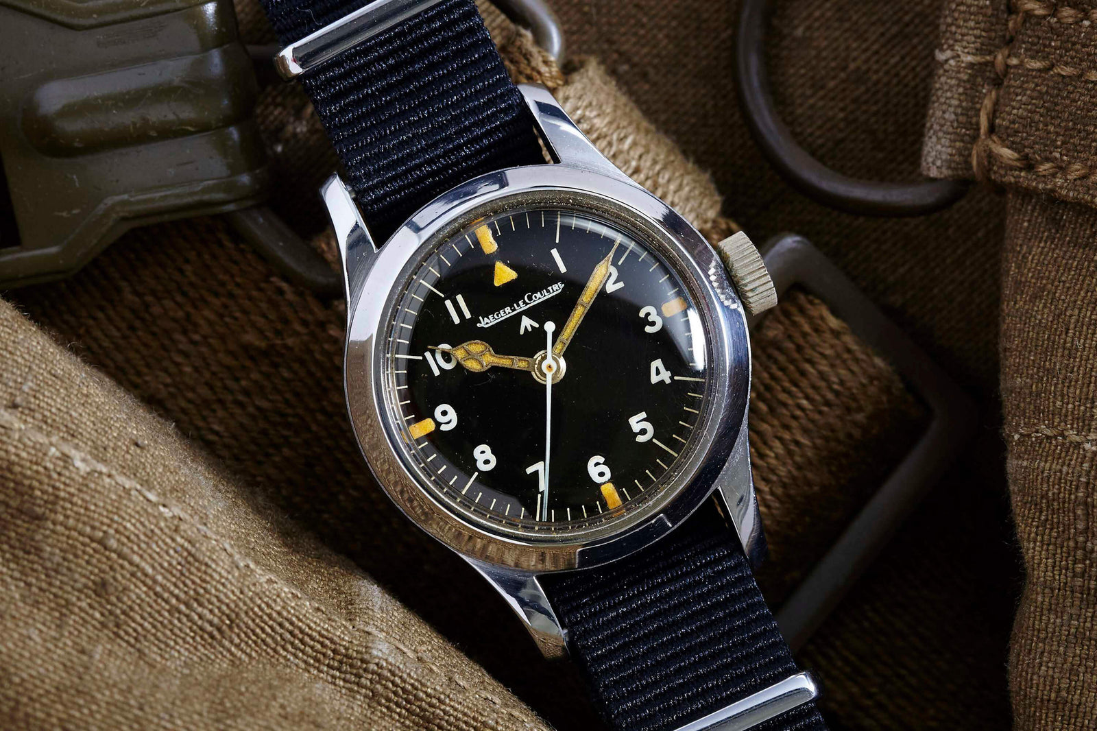 The Jaeger LeCoultre JLC Mark XI Pilot's watch has a black dial with white numerals, yellow accents, anti-magnetic properties, and a black fabric strap. It is displayed on brown textured fabric with metal hardware.