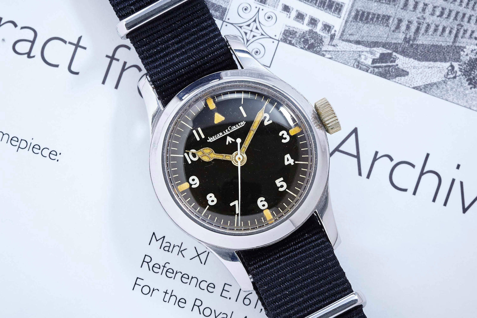 A Jaeger LeCoultre JLC Mark XI pilot's watch with a black dial, luminous numerals, and a black nylon strap, known for its anti-magnetic features, sits on a document with partially visible printed text.