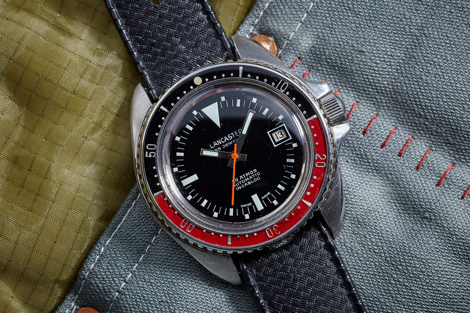 A close-up of the Lancaster 844 Diver by Lancaster, featuring a black dial, red and black bezel, and textured strap. It rests on green and gray fabric with orange stitching, displaying the date as the 13th.
