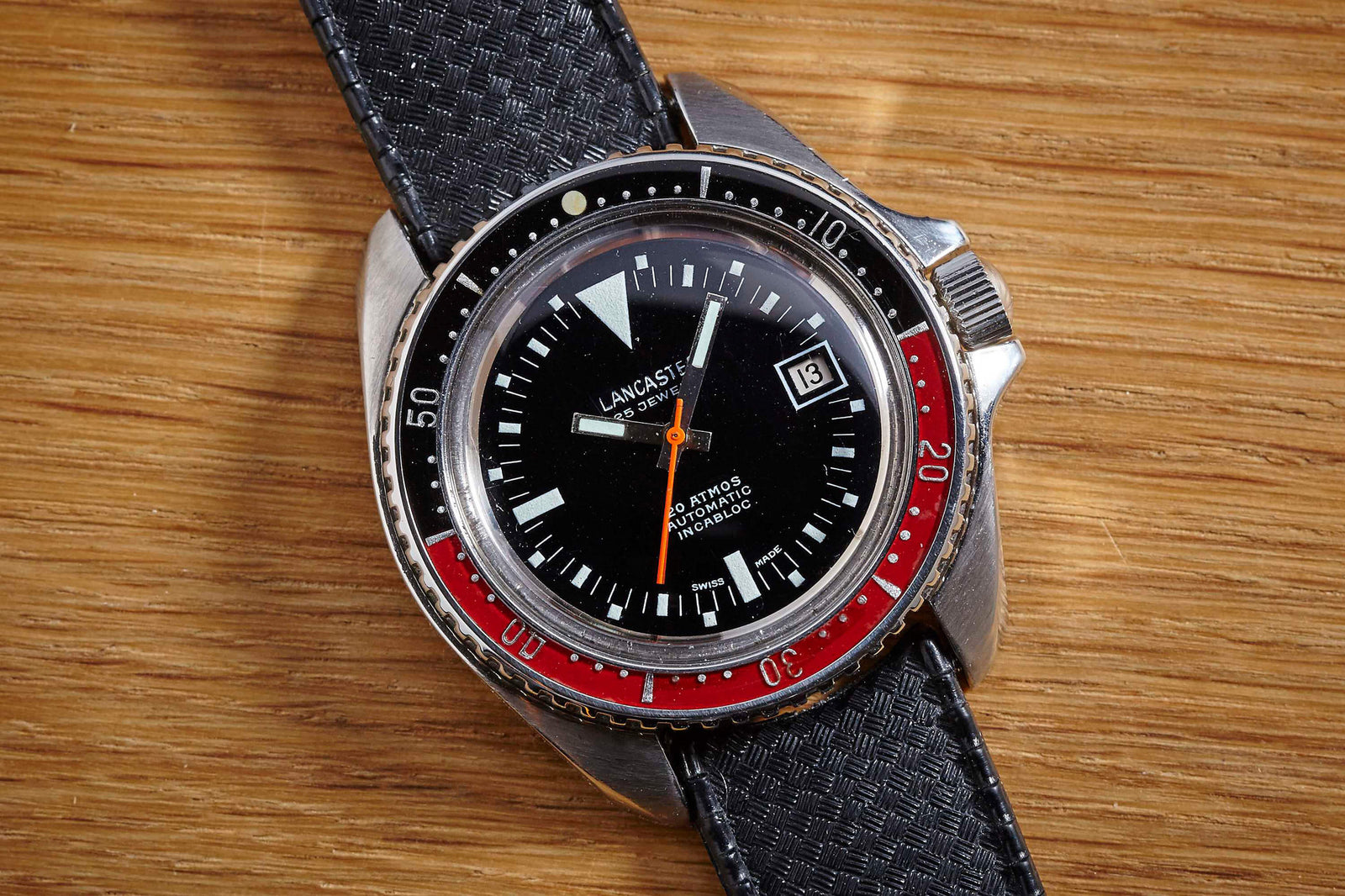 A close-up of the Lancaster 844 Diver by Lancaster, featuring a black dial, white and orange markers, red and black rotating bezel, date display, and textured strap resting on a wooden surface.