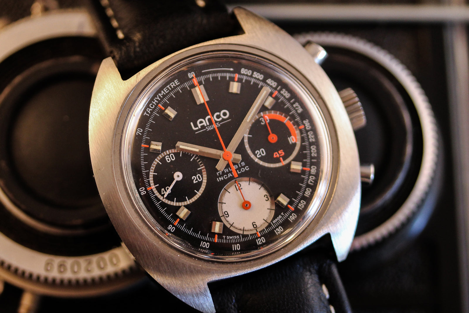 A close-up of the Lanco Chronograph by Lanco, featuring a black dial with three subdials, orange and white accents, and a black leather strap on a dark textured background.