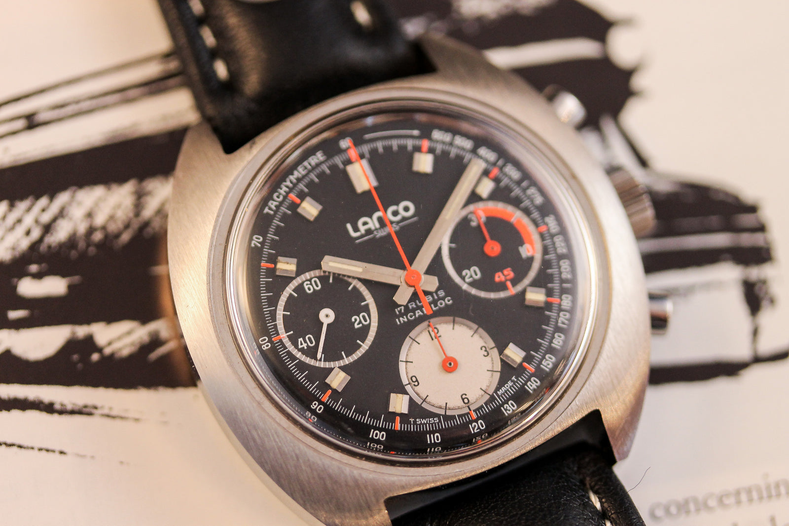 Close-up of the Lanco Chronograph, a vintage watch by Lanco with a black dial, three subdials, Valjoux 7736 movement, silver case, tachymeter scale, and black leather strap on a monochrome background.