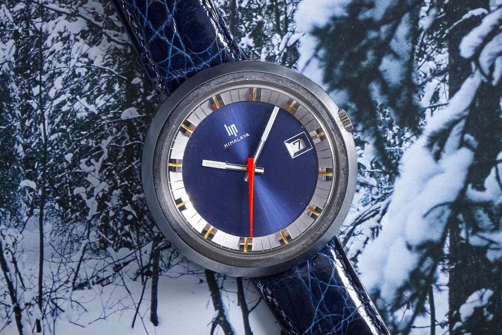 A close-up of the Lip Himalaya wristwatch by Lip, featuring a blue face, red second hand, and blue leather strap against a snowy, black-and-white forest backdrop. The date window displays seven—a bold piece from the French brand.