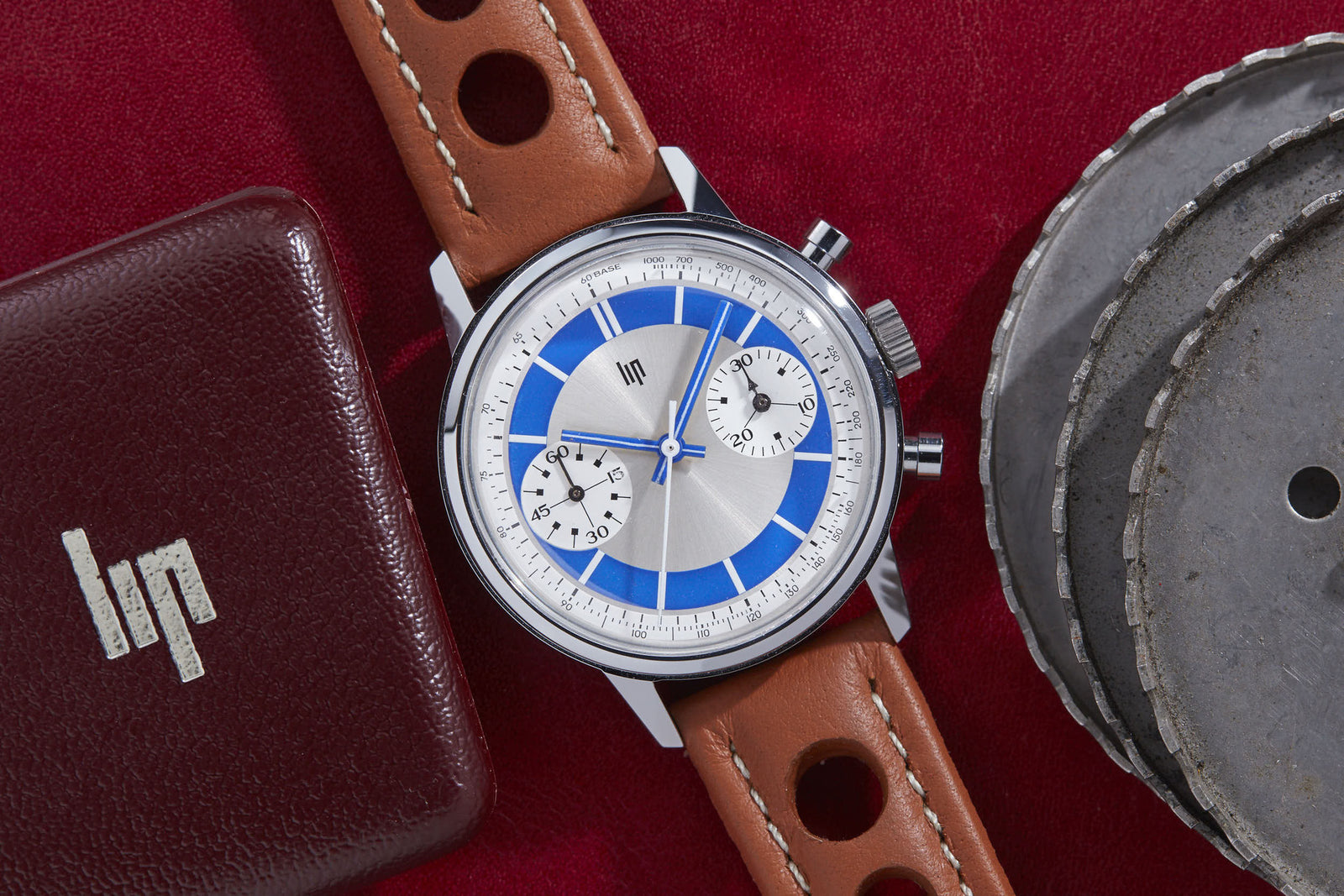 The Lip "Paul Newman" Chronograph, featuring a brown leather strap and blue-and-silver dial, rests on a red surface beside a burgundy leather case with a white Lip logo and two stacked metal discs.