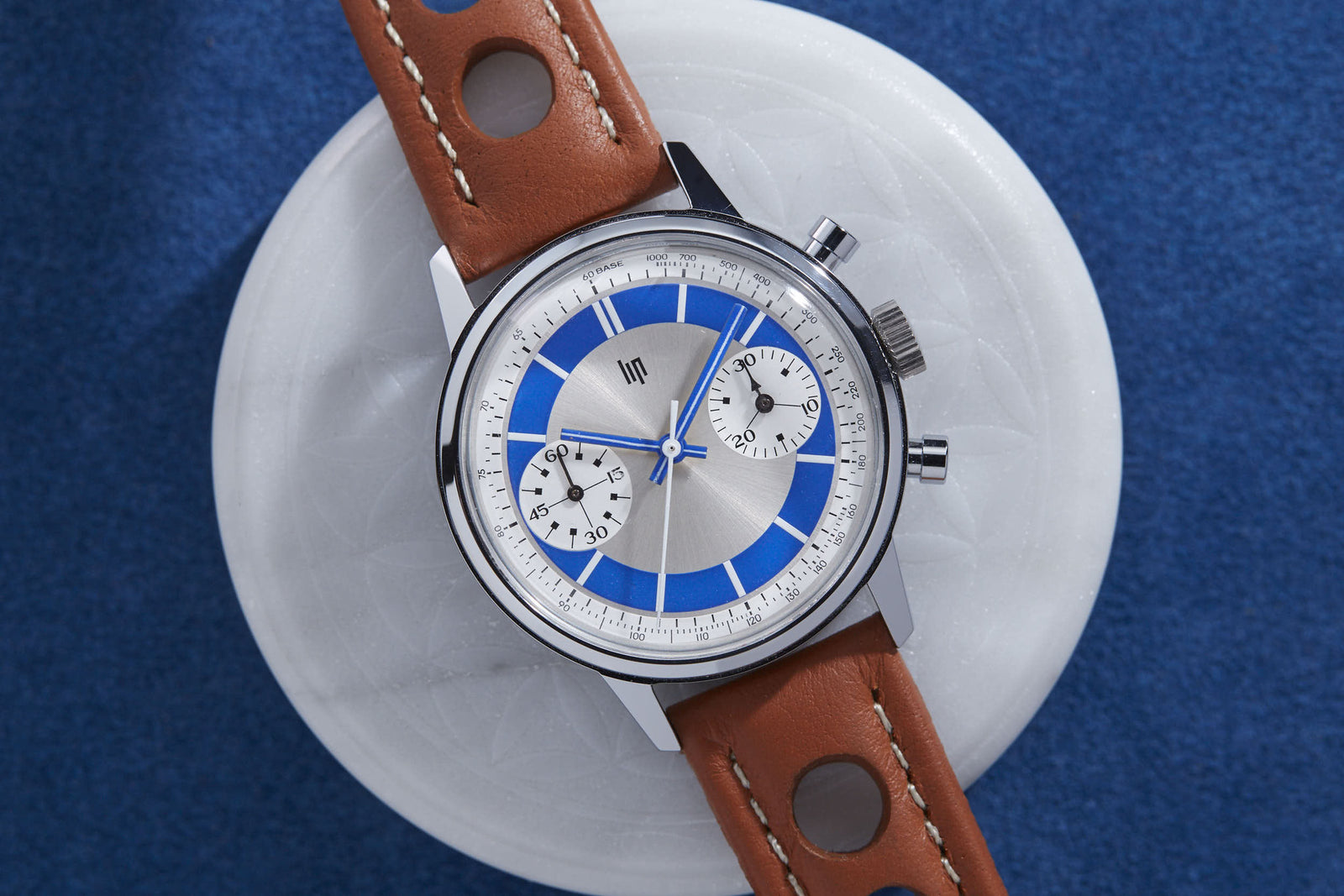 The Lip "Paul Newman" Chronograph, featuring a silver case, blue and white details, brown perforated leather strap, and two subdials, is displayed on a round white object set against a blue background.