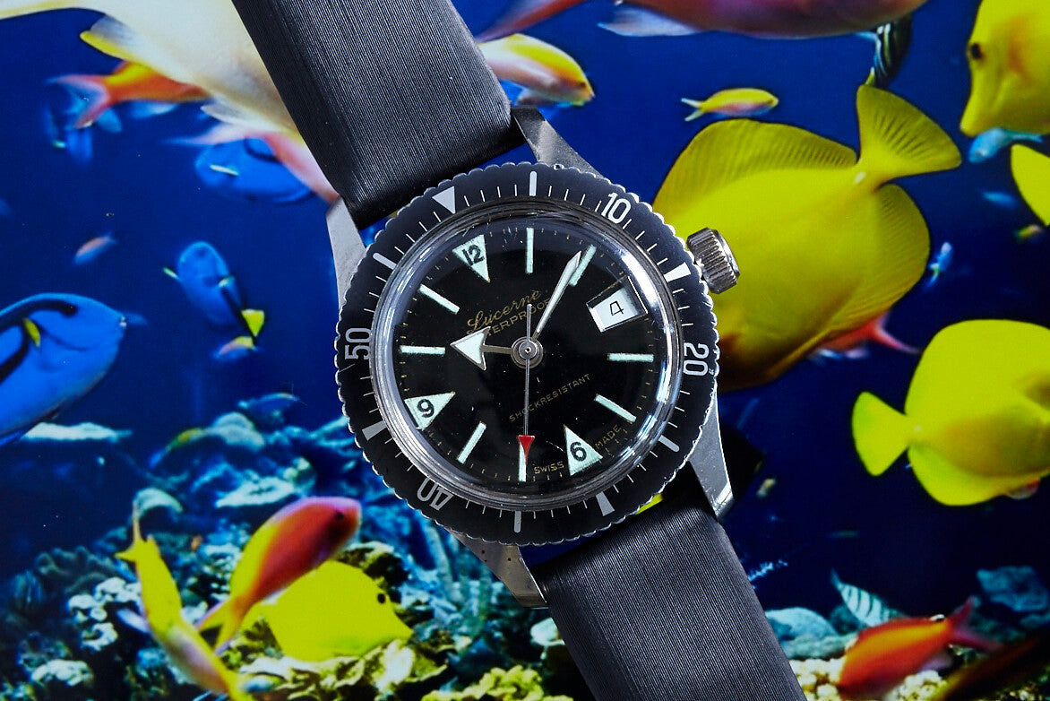 The Lucerne Lady Diver by Lucerne is a vintage ladies' black wristwatch with a metal bezel and dark strap, shown against a vibrant underwater backdrop with yellow, blue, and red fish among coral reefs.