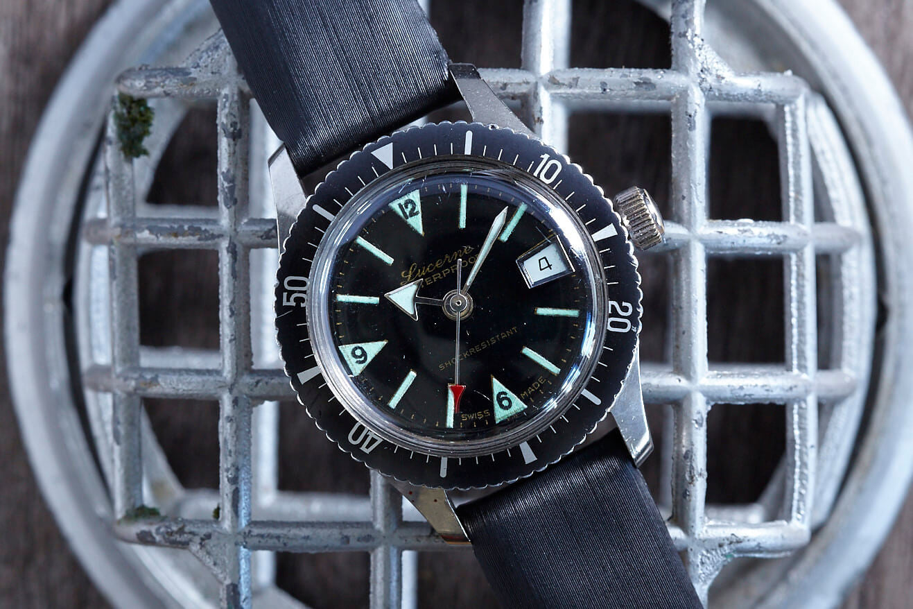 The Lucerne Lady Diver by Lucerne, a vintage manual-winder, rests on a metal grid. It features a black strap and rotating bezel, black face with luminescent hands, bold numerals at 3, 6, 9, and 12, plus a date window at 3 o’clock.
