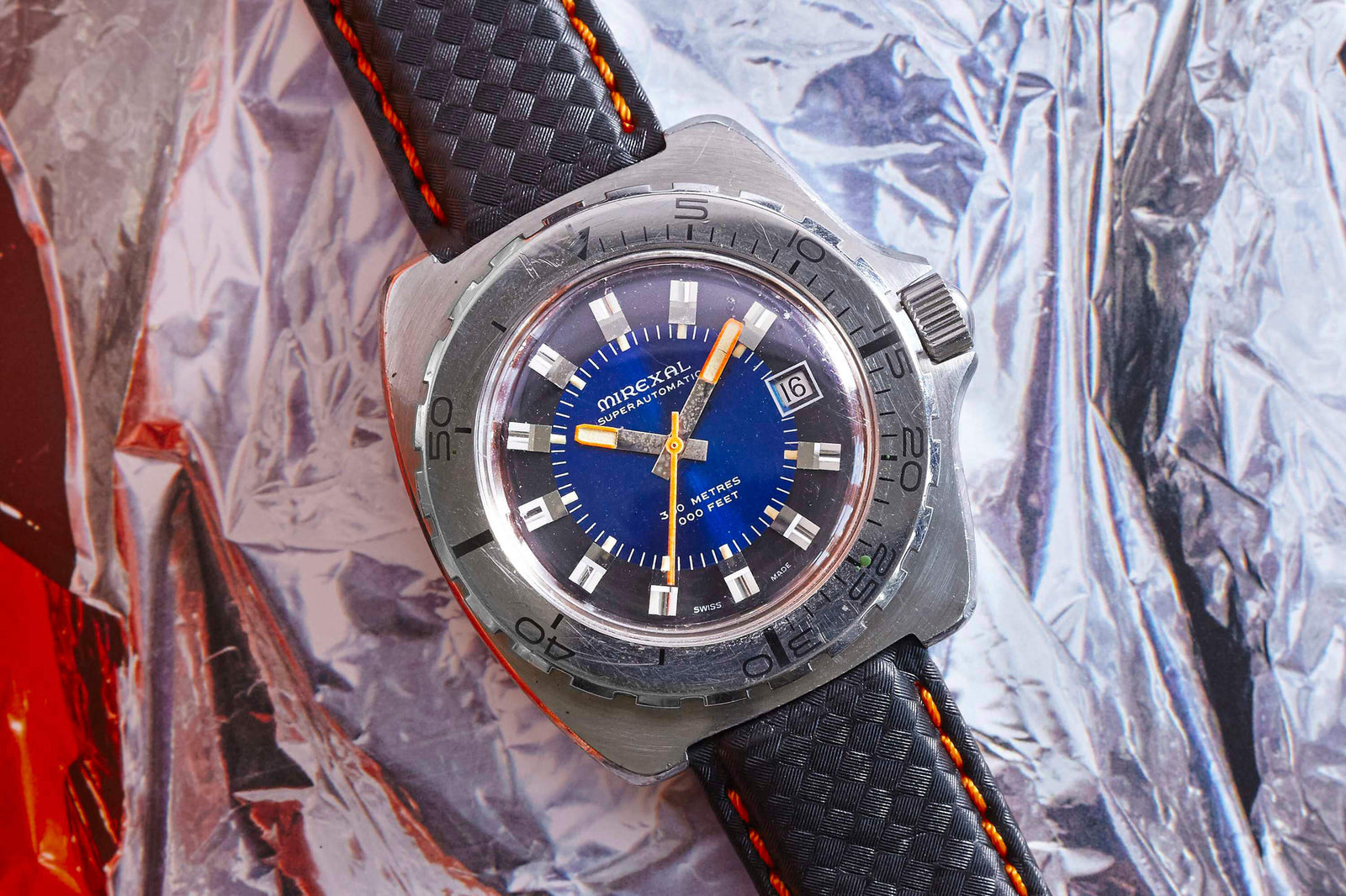 The Mirexal SuperAutomatic wristwatch, a vintage-inspired silver timepiece with a blue dial, black strap with orange stitching, and date at 3 o’clock, rests on crinkled reflective metal.