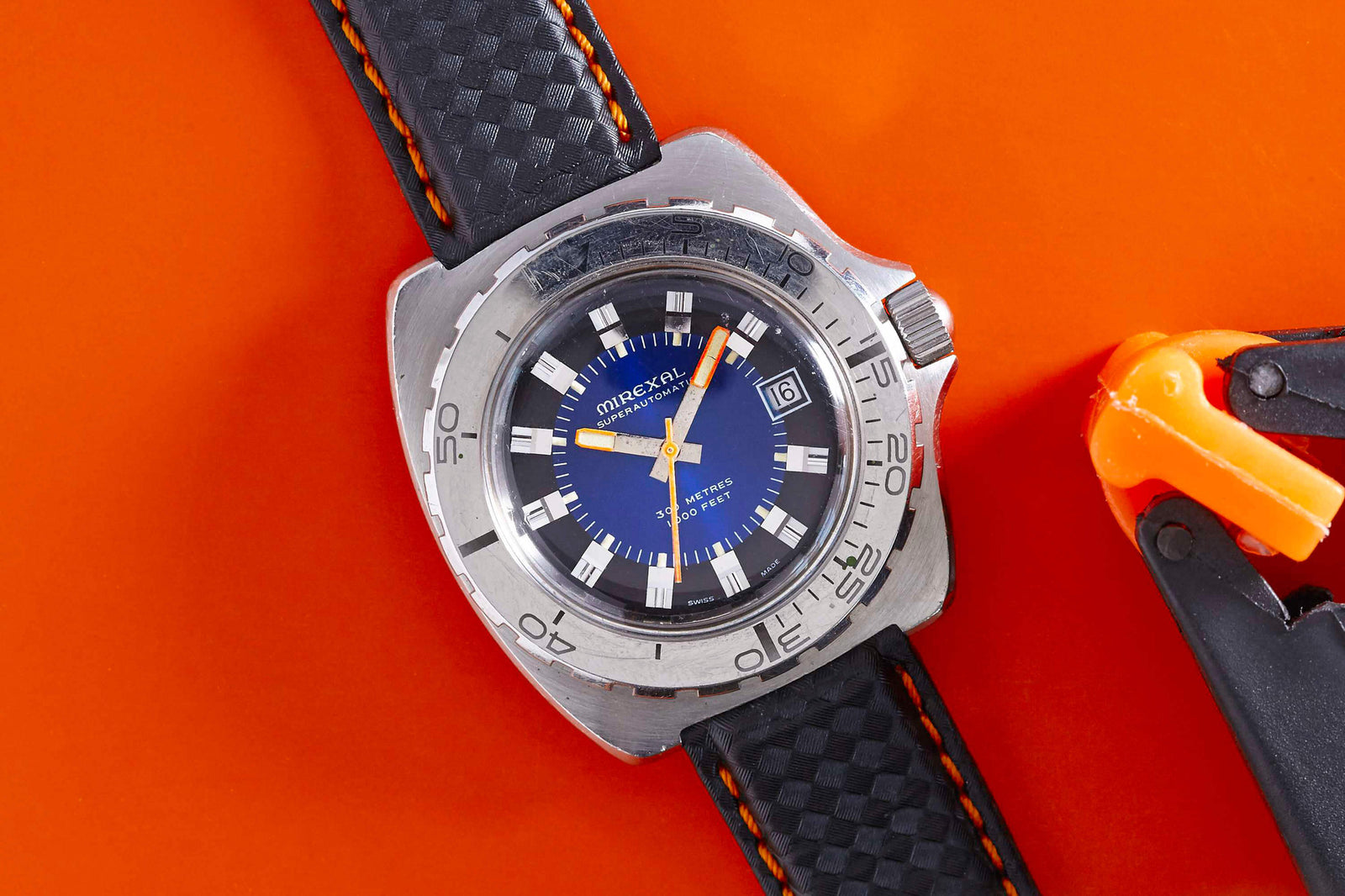 The Mirexal SuperAutomatic by Mirexal is a vintage-inspired dive watch with a blue dial, silver bezel, black band with orange stitching, white hour markers, and a date display—all set against an orange surface for a classic 1960s look.