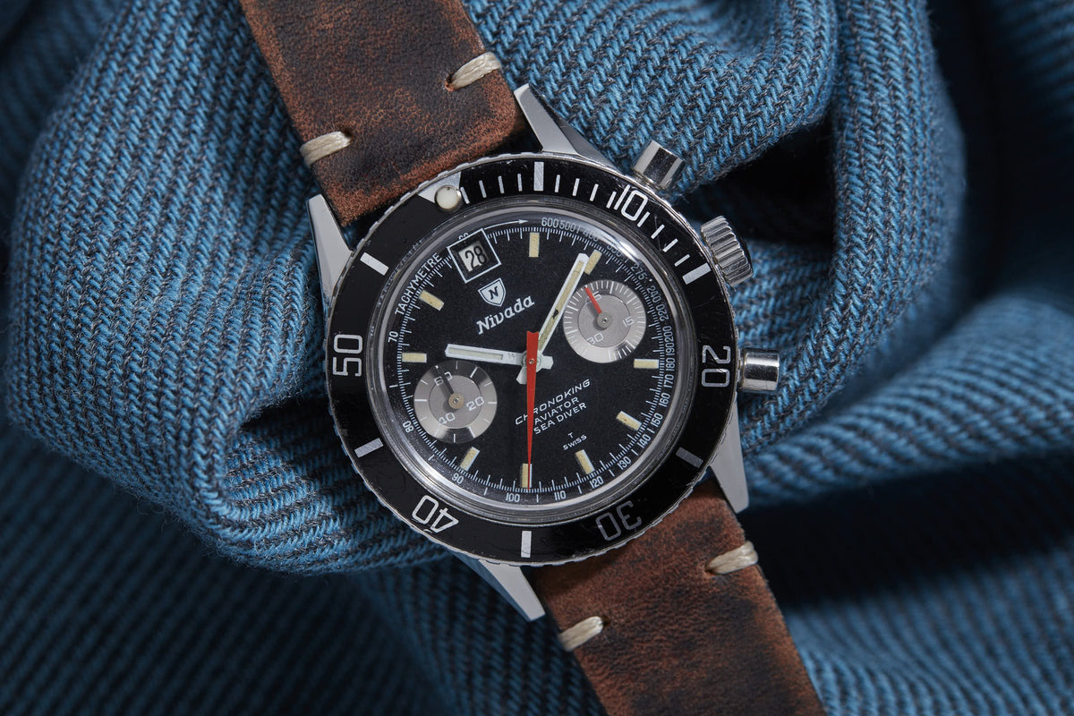 Nivada Chronoking Date watch with black dial, chronograph subdials, date window, and brown leather strap on blue fabric backg
