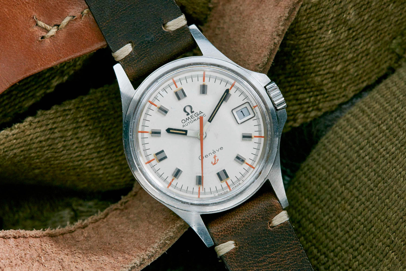 An Omega Genève Admiralty wristwatch with a white dial, black and orange markers, and a brown leather strap rests on textured green fabric. The classic Omega watch displays the date with "Automatic" and the Omega logo on the face.