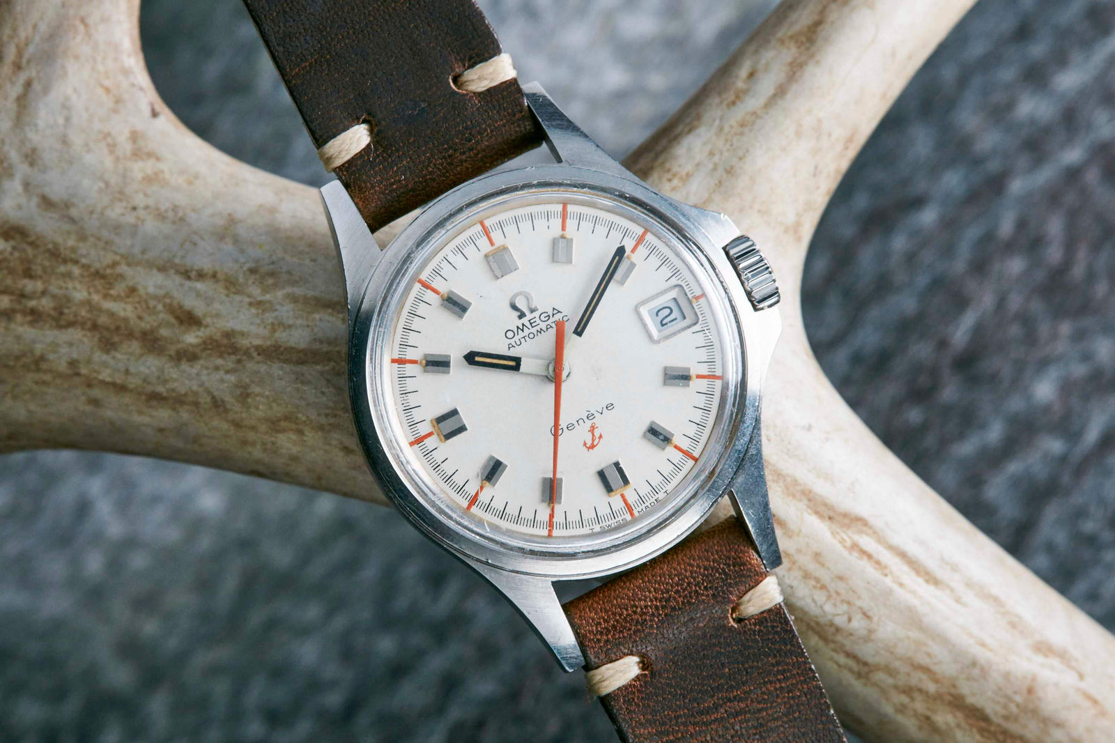 An Omega Genève Admiralty wristwatch with a silver dial, date window at 3 o’clock, orange accents, and a brown leather strap rests on a light-colored antler. This classic Omega timepiece exudes timeless style.