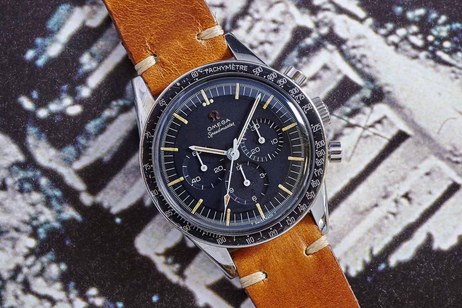 The Omega Speedmaster "Ed White" by Omega is shown close up, featuring a black dial, chronograph subdials, and a brown leather strap on a black and white textured background. Detailed description coming soon!.