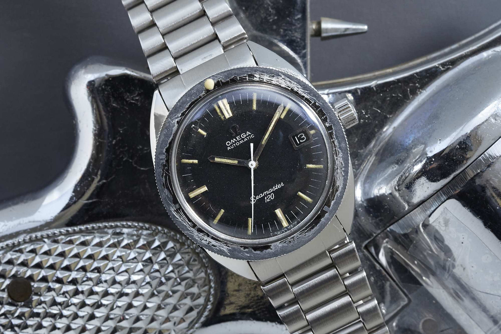 An Omega Seamaster 120 dive watch with a black dial and metal band sits on a shiny surface, showing the time at 5:36 and the date as 13.