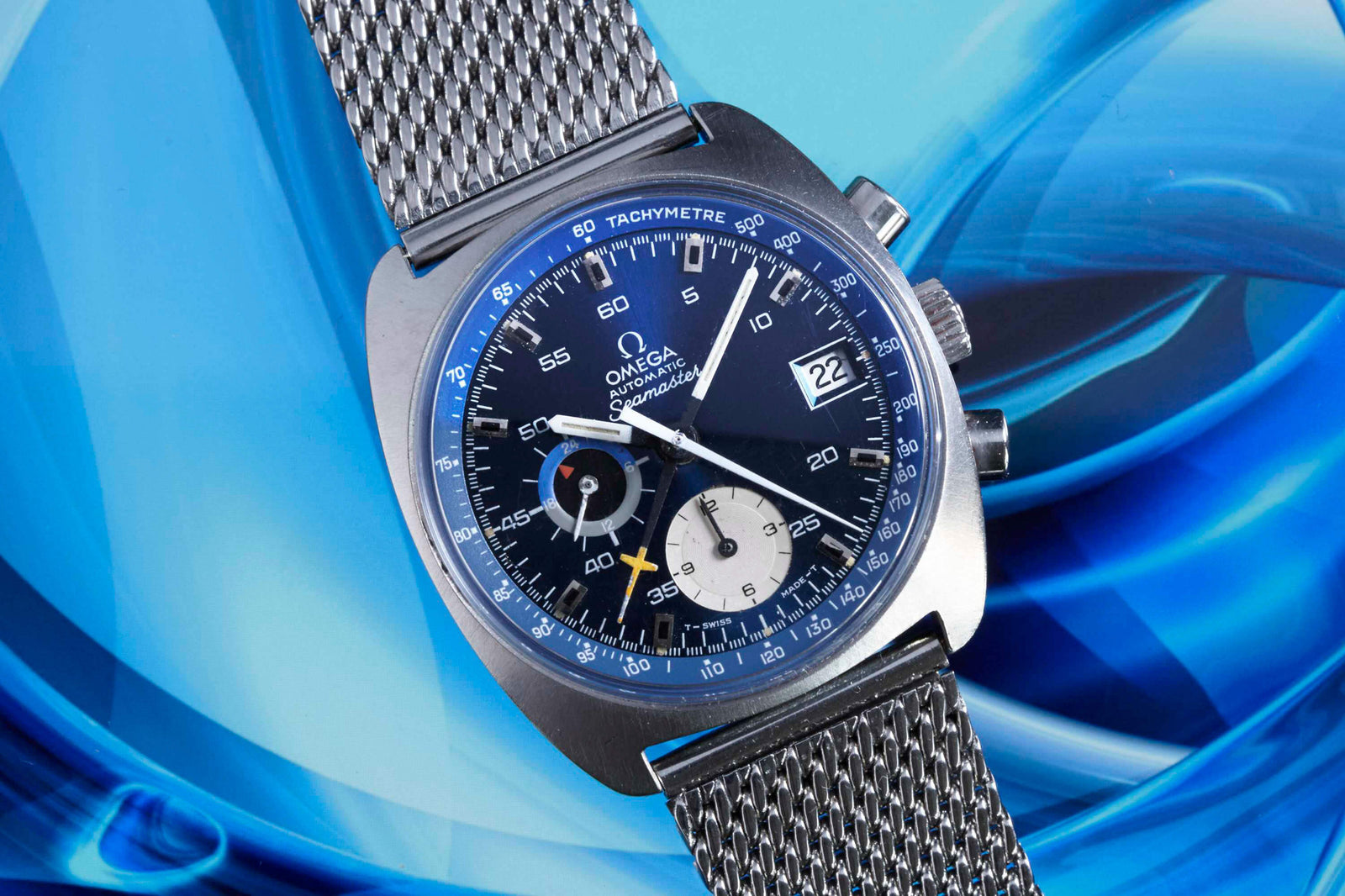 A close-up of the Omega Seamaster Chronograph Automatic by Omega, featuring a blue dial, silver mesh strap, and chronograph functions, set against a vibrant blue background—ideal for vintage watch enthusiasts.