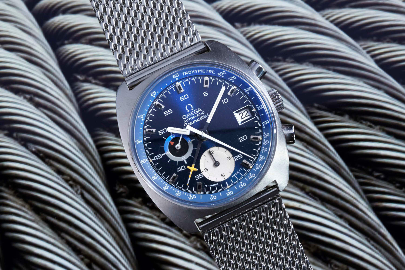 A close-up of the Omega Seamaster Chronograph Automatic by Omega, featuring a blue and black dial, silver mesh strap, and tachymeter scale, with the date at 3 o’clock, set atop twisted metal cables—a timeless piece for vintage watch lovers.