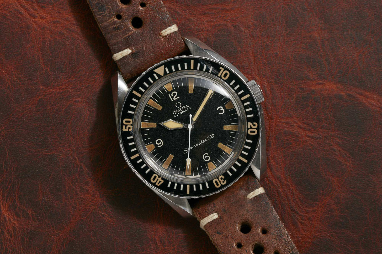 The Omega Seamaster 300 by Omega, featuring a black dial, cream hour markers, and a brown leather strap, rests on textured leather. More details coming soon!.