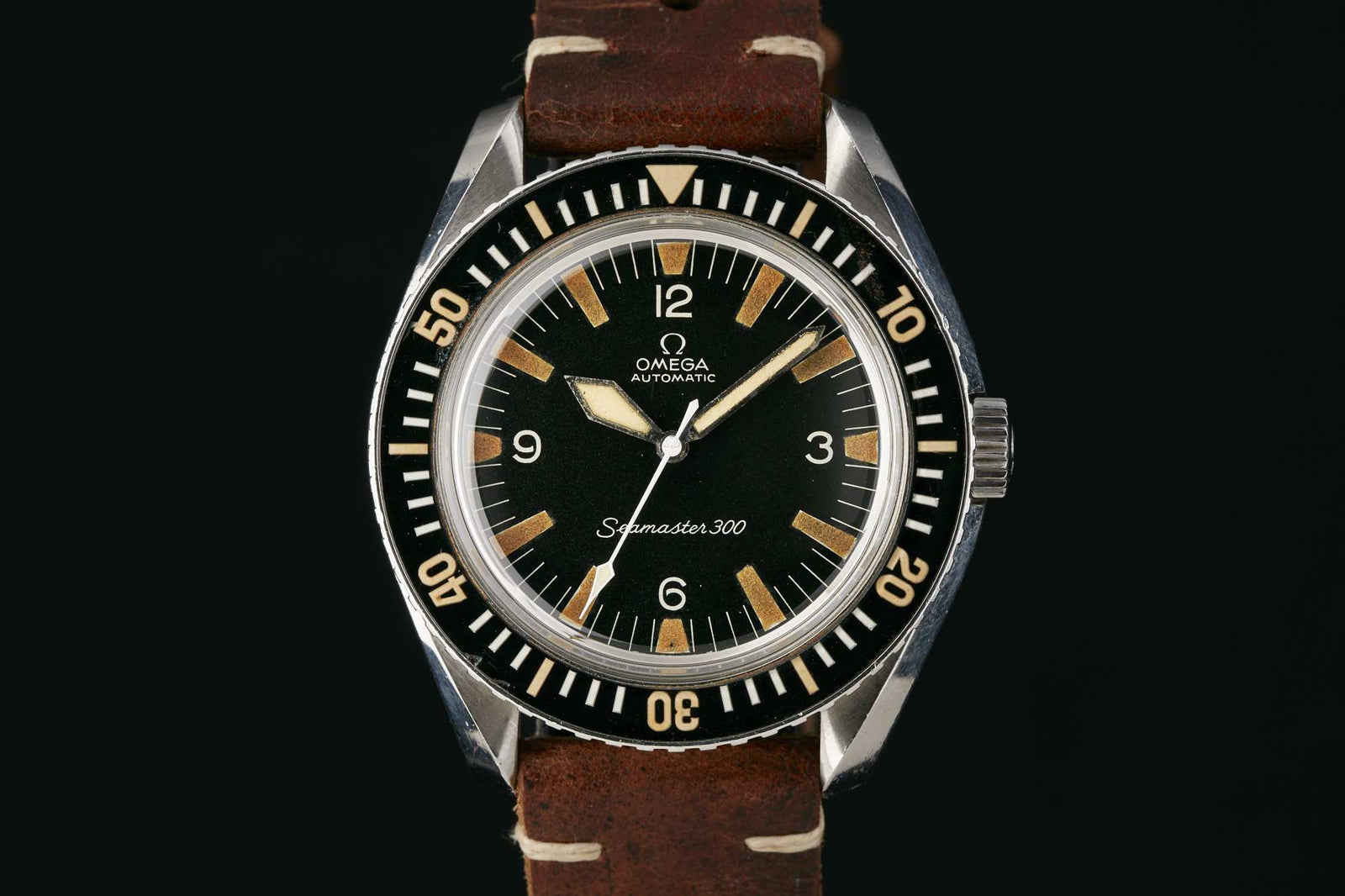 The Omega Seamaster 300 by Omega features a black dial with bold white numerals and indexes, set in a stainless steel case with a rotating bezel and brown leather strap. Detailed description coming soon! Shown on a black background.