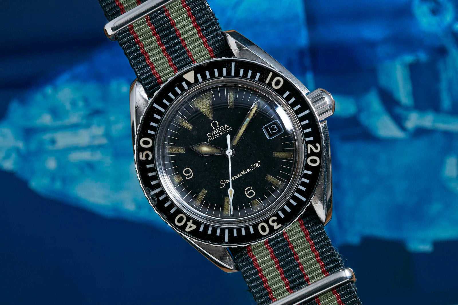 A close-up of the Omega Seamaster 300 by Omega, featuring a black dial, white markers, date at 3 o’clock, and a striped NATO strap against a blurred blue background. This waterproof sports watch combines style and function.