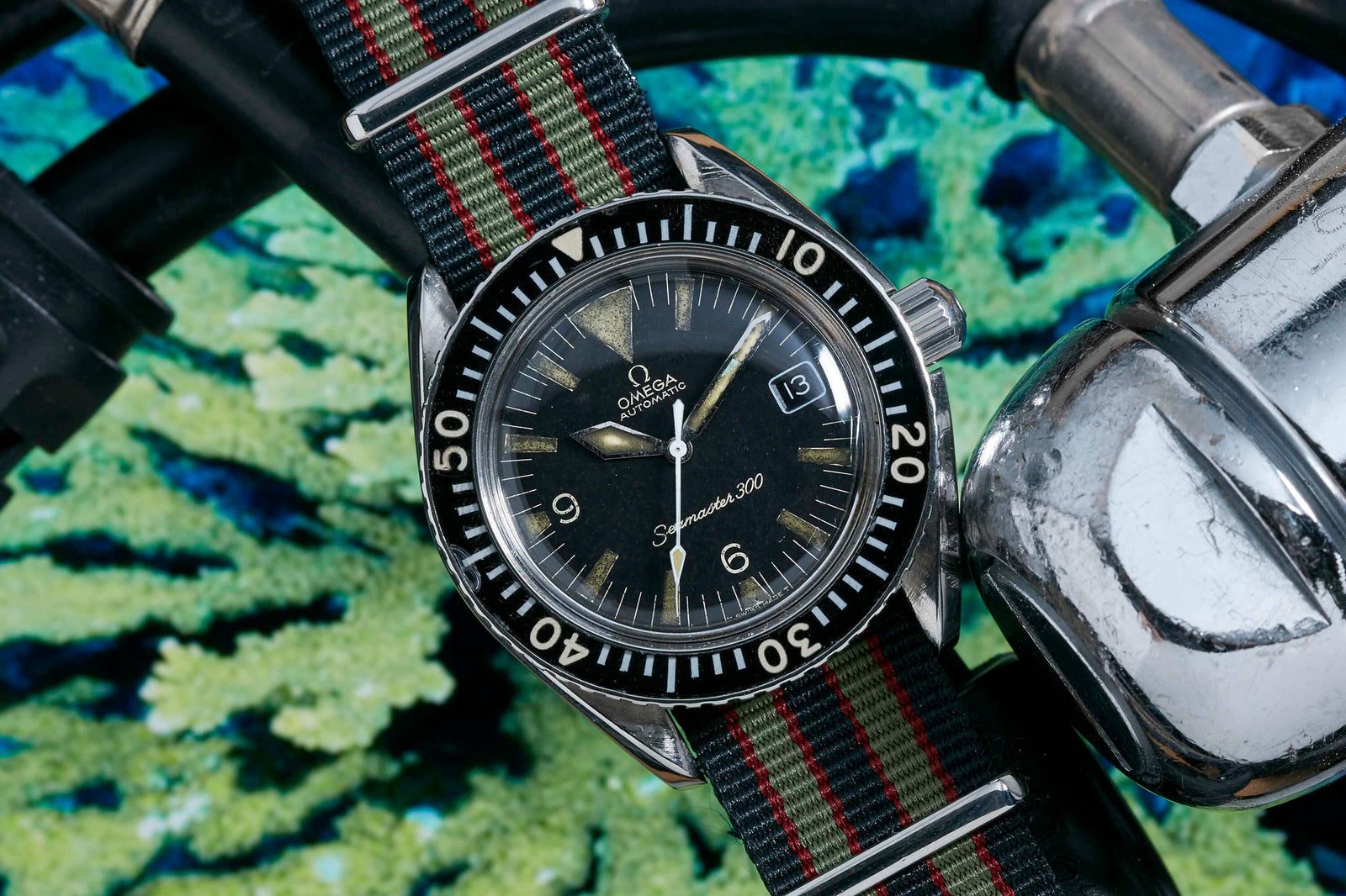 A close-up of the Omega Seamaster 300 by Omega, a waterproof sports watch, features a striped black and red nylon strap beside diving gear with underwater coral in the background.