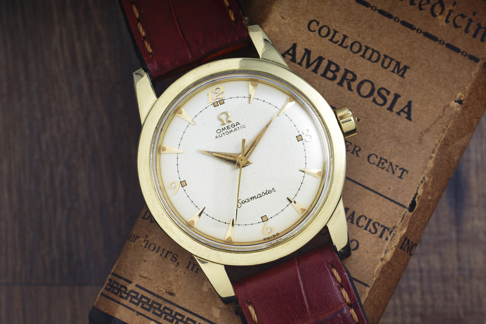 An Omega Seamaster Automatic - Yellow Gold Filled by Omega with a brown leather strap and gold hands on a white dial rests atop the book Colloidum Ambrosia.