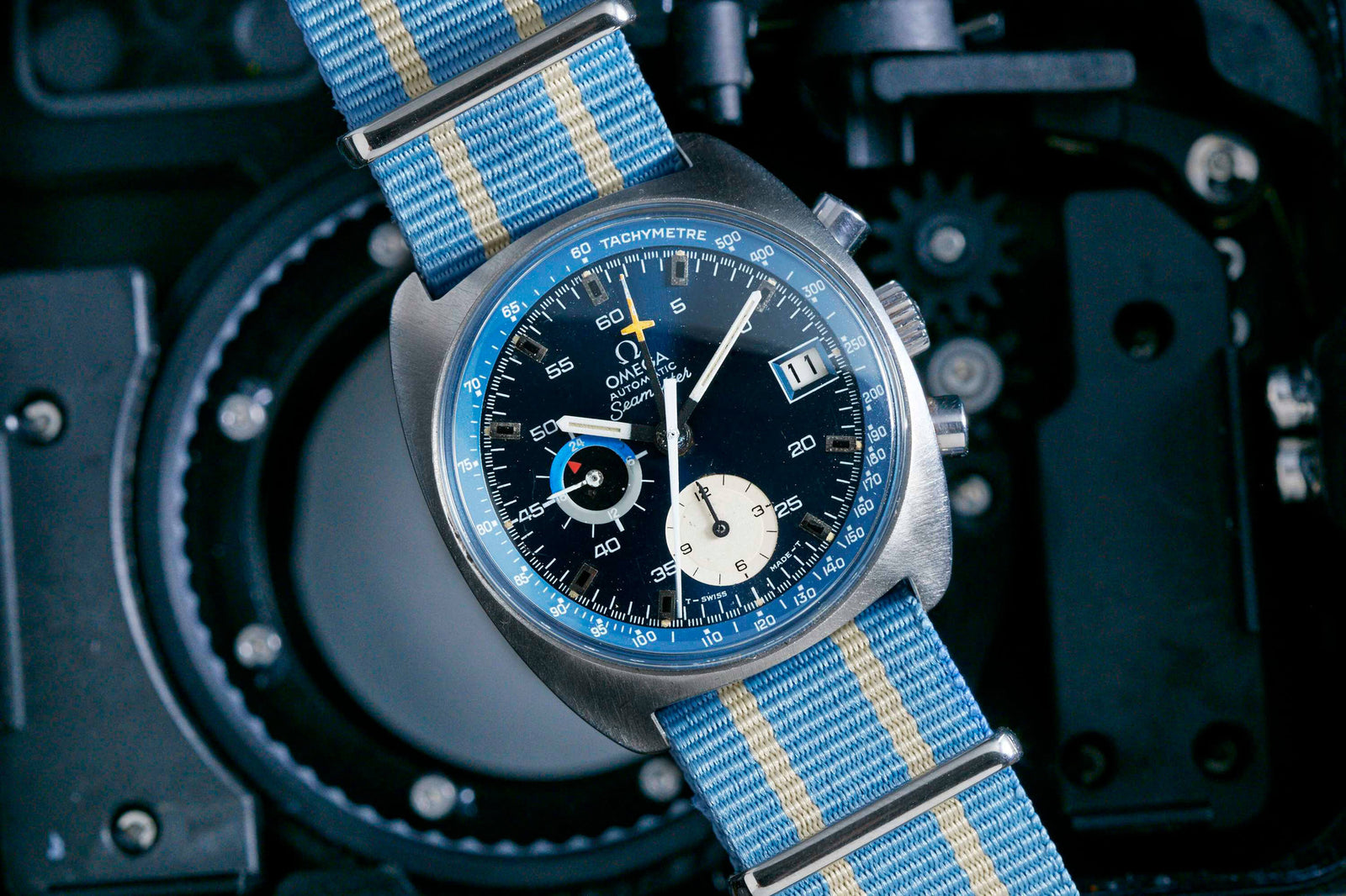 A close-up of an Omega Seamaster Automatic Chronograph wristwatch with a blue, white, and beige striped strap, blue tachymeter bezel, black dial, and two subdials set against a dark mechanical background.