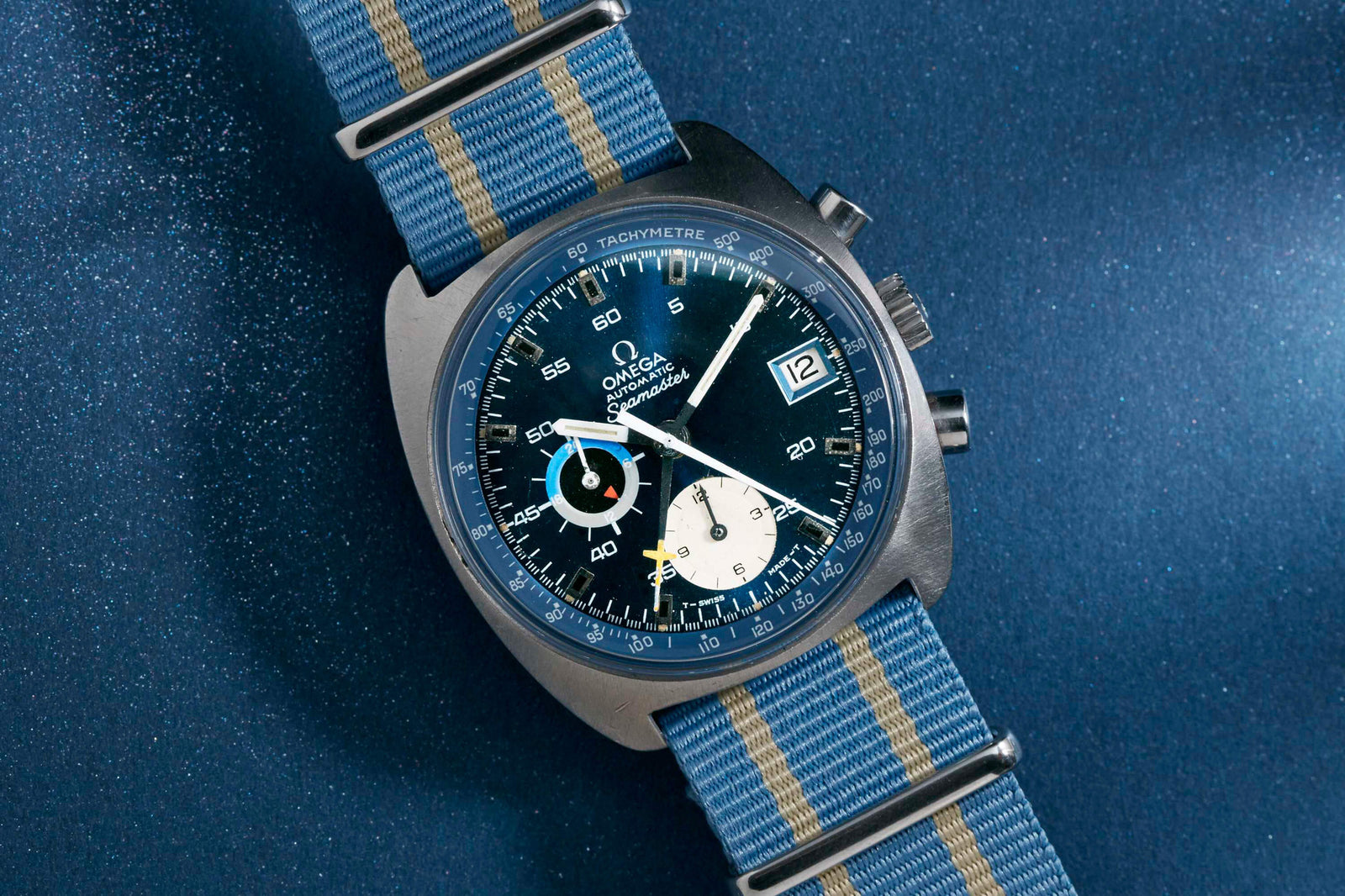 An Omega Seamaster Automatic Chronograph by Omega, featuring a blue dial, tachymeter bezel, white and yellow accents, automatic movement, and a blue-striped NATO strap, displayed against a sparkling blue background.