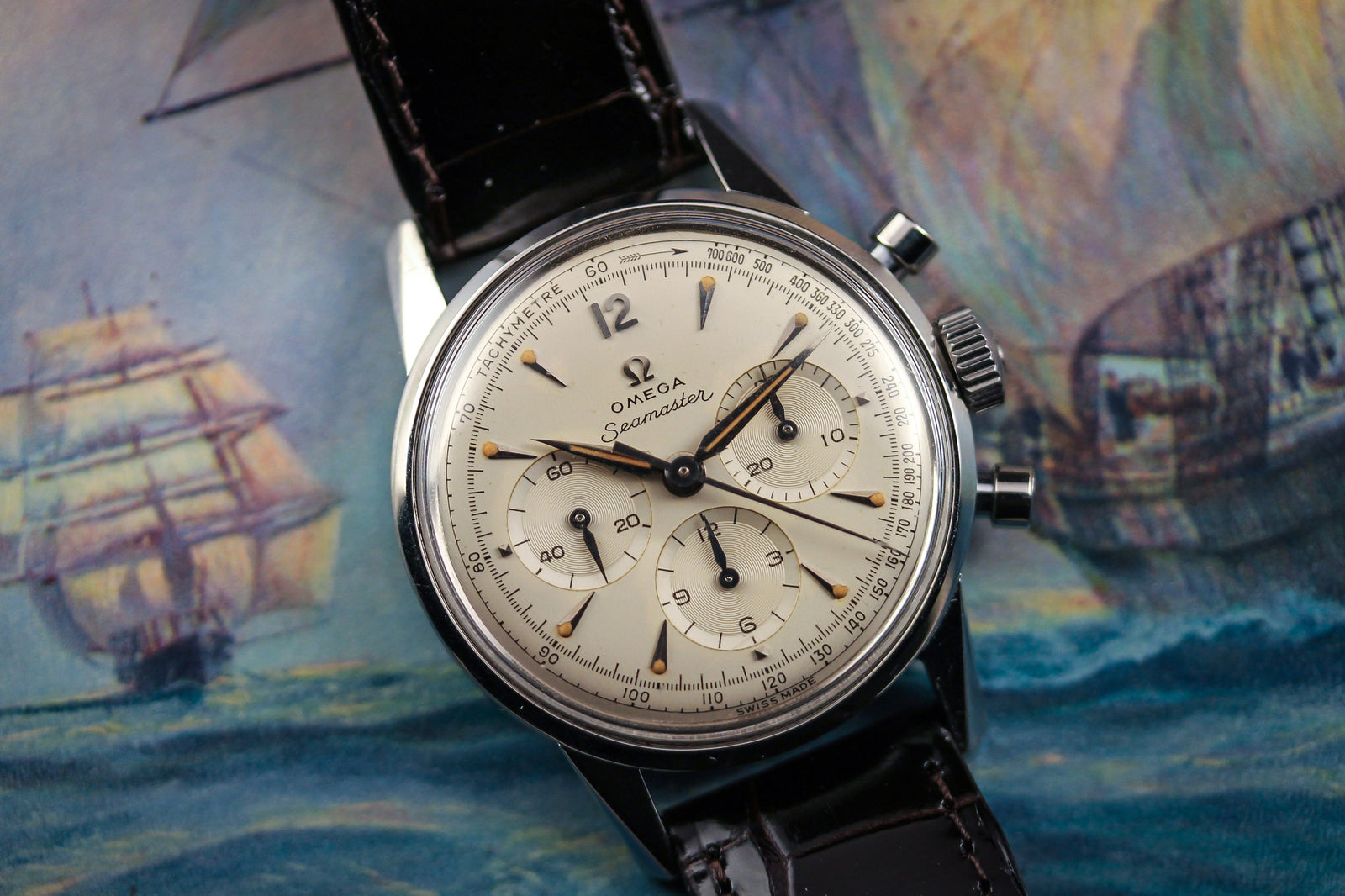 The Omega Seamaster Chronograph Calibre .321 - 1960 by Omega features a cream dial, three subdials, and a black leather strap, displayed on nautical-themed art with sailing ships to showcase its classic movement.